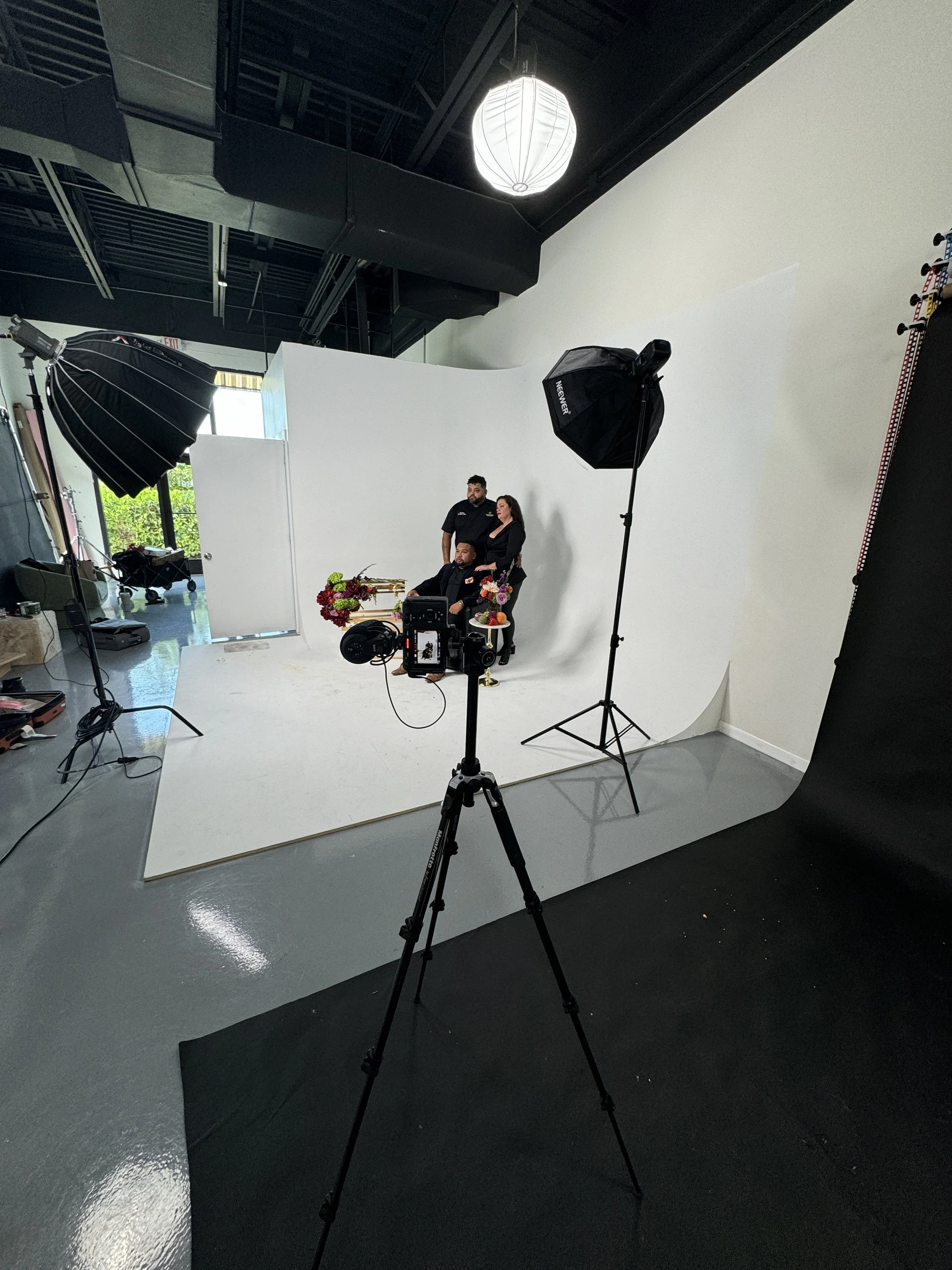 Photo of a photo studio with a white backdrop, studio lights, and three people posing for a photograph, with flowers on a table in front of them.
