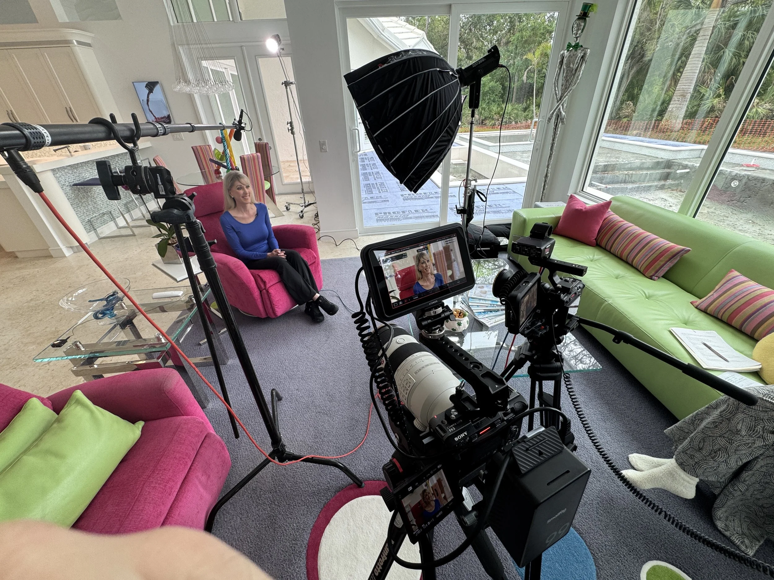 A woman sitting on a pink sofa being filmed in a home studio with colorful couches and large windows showing outdoor greenery.