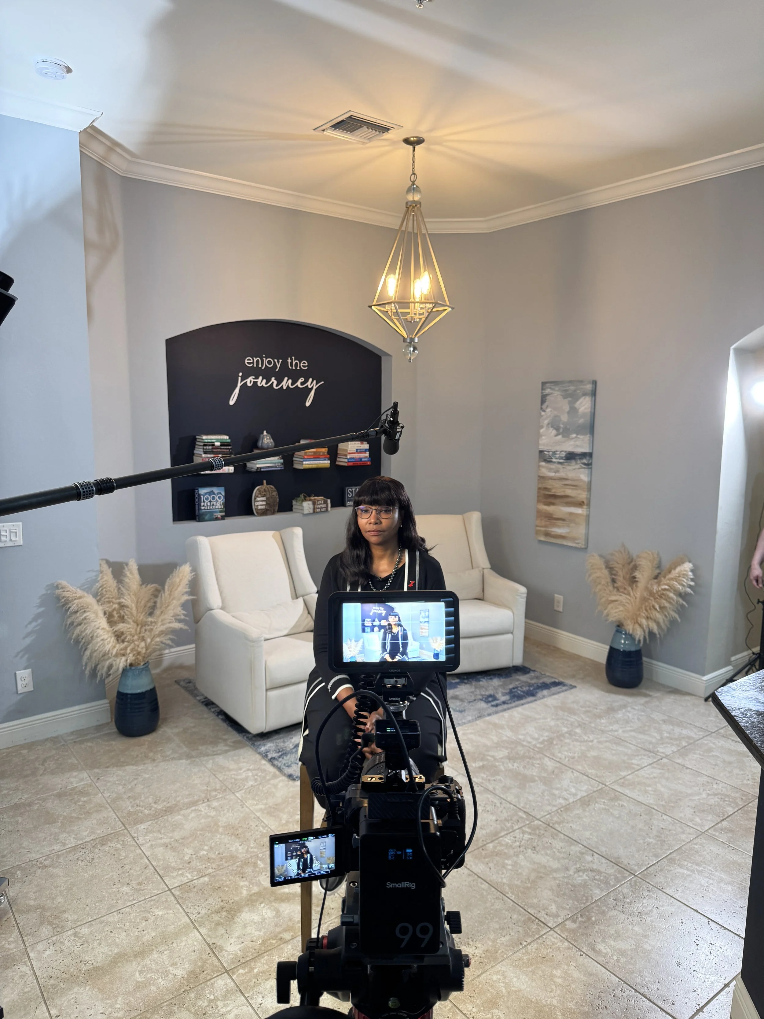 A woman with glasses and dark hair, sitting on a chair, being filmed inside a living room with light gray walls, a white couch, two blue vases with pampas grass, and framed artwork, under a modern geometric chandelier.