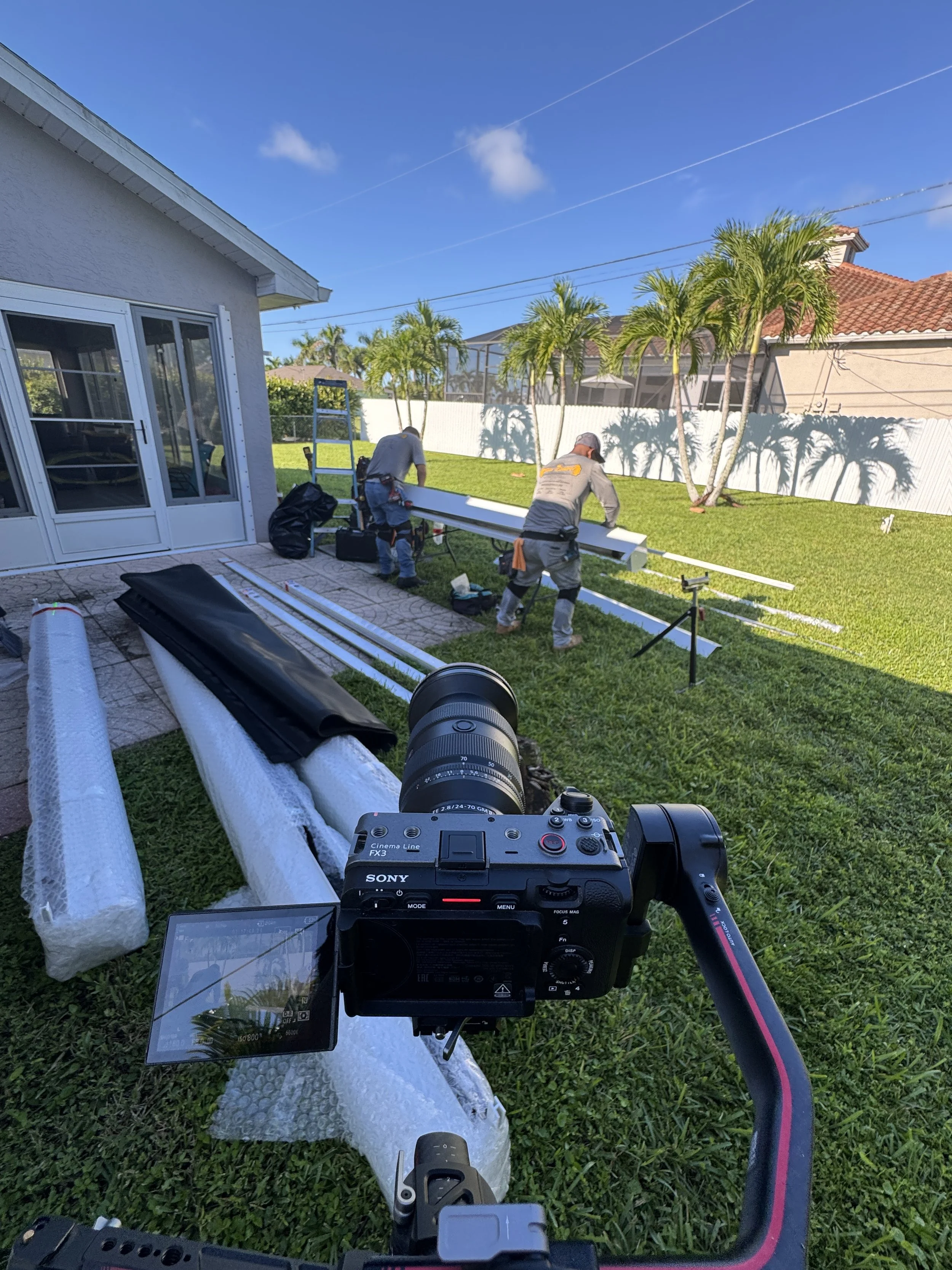 View from a camera mounted on a drone overlooking two workers installing siding or trim on the exterior of a house in a backyard with green grass, palm trees, and a white fence.