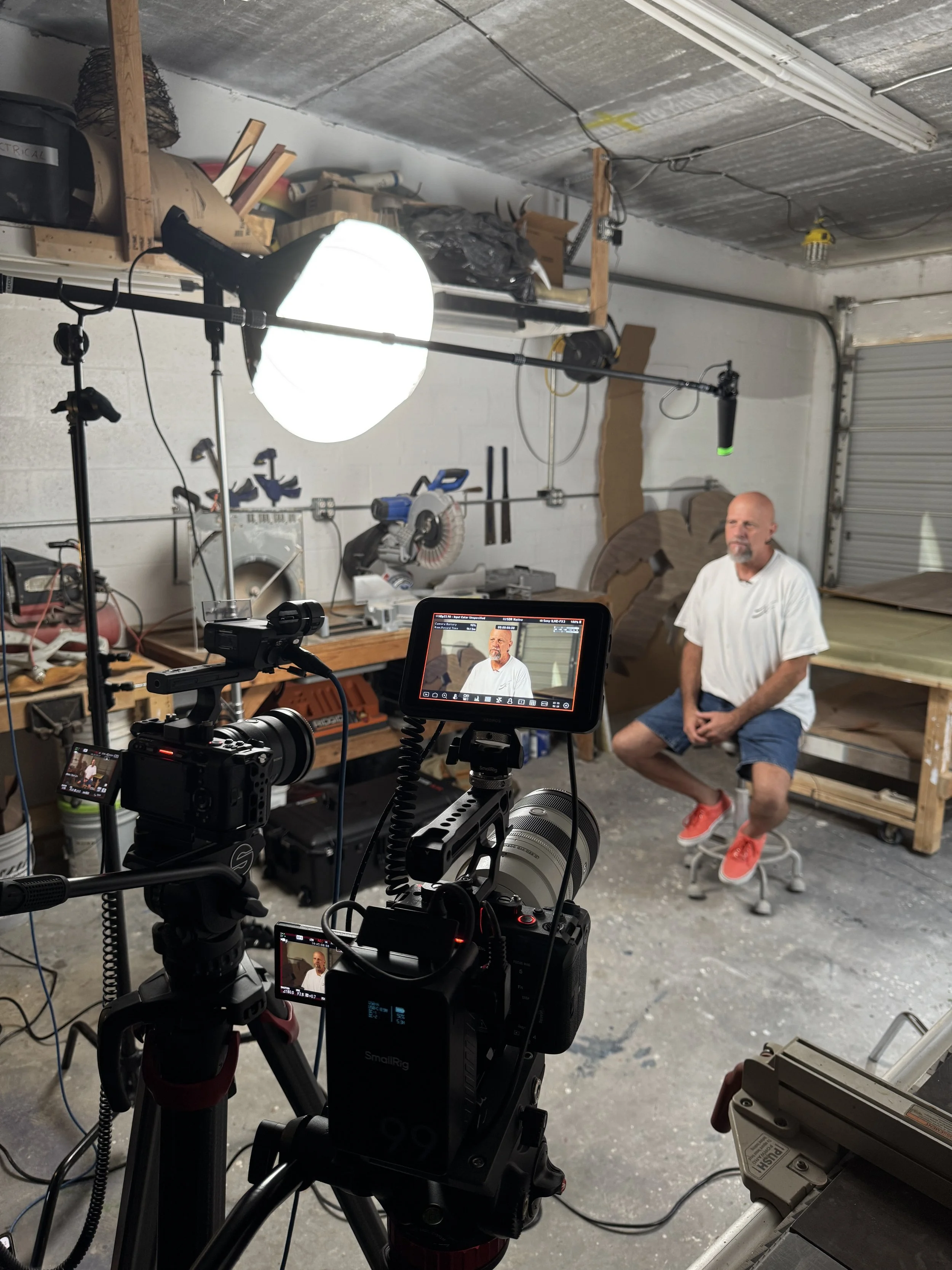 A man with a bald head and beard is sitting on a stool in a workshop or studio with a concrete floor and storage shelves. There is photography or video filming equipment, including cameras, lights, and monitors, set up around him. The man is wearing 
