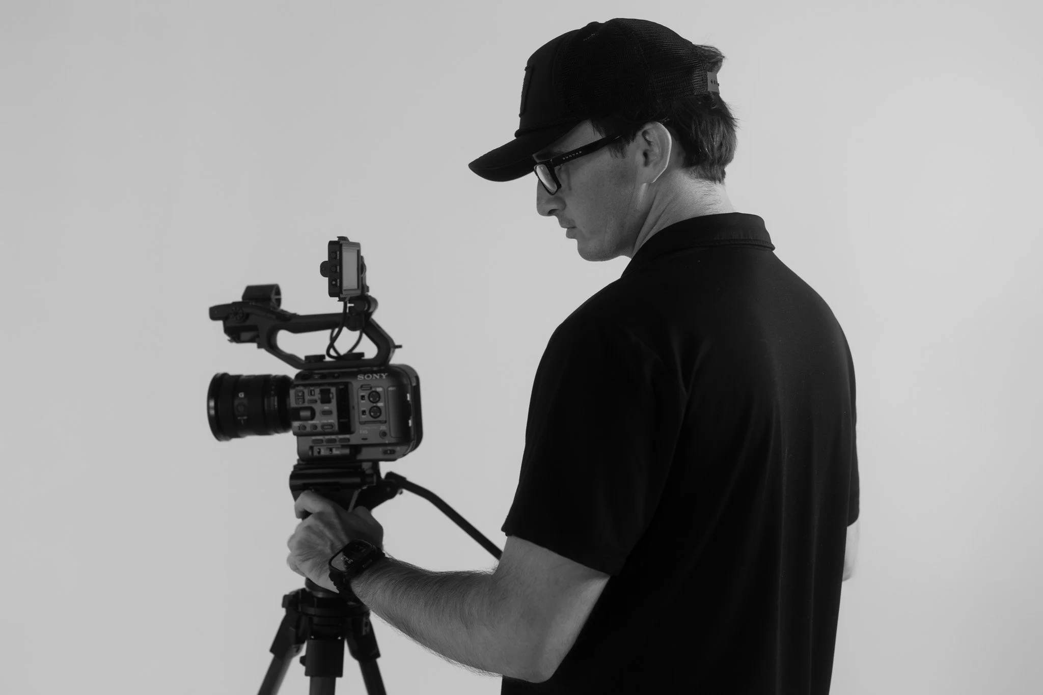 A man in a black shirt and cap, wearing glasses, adjusting a professional video camera mounted on a tripod.