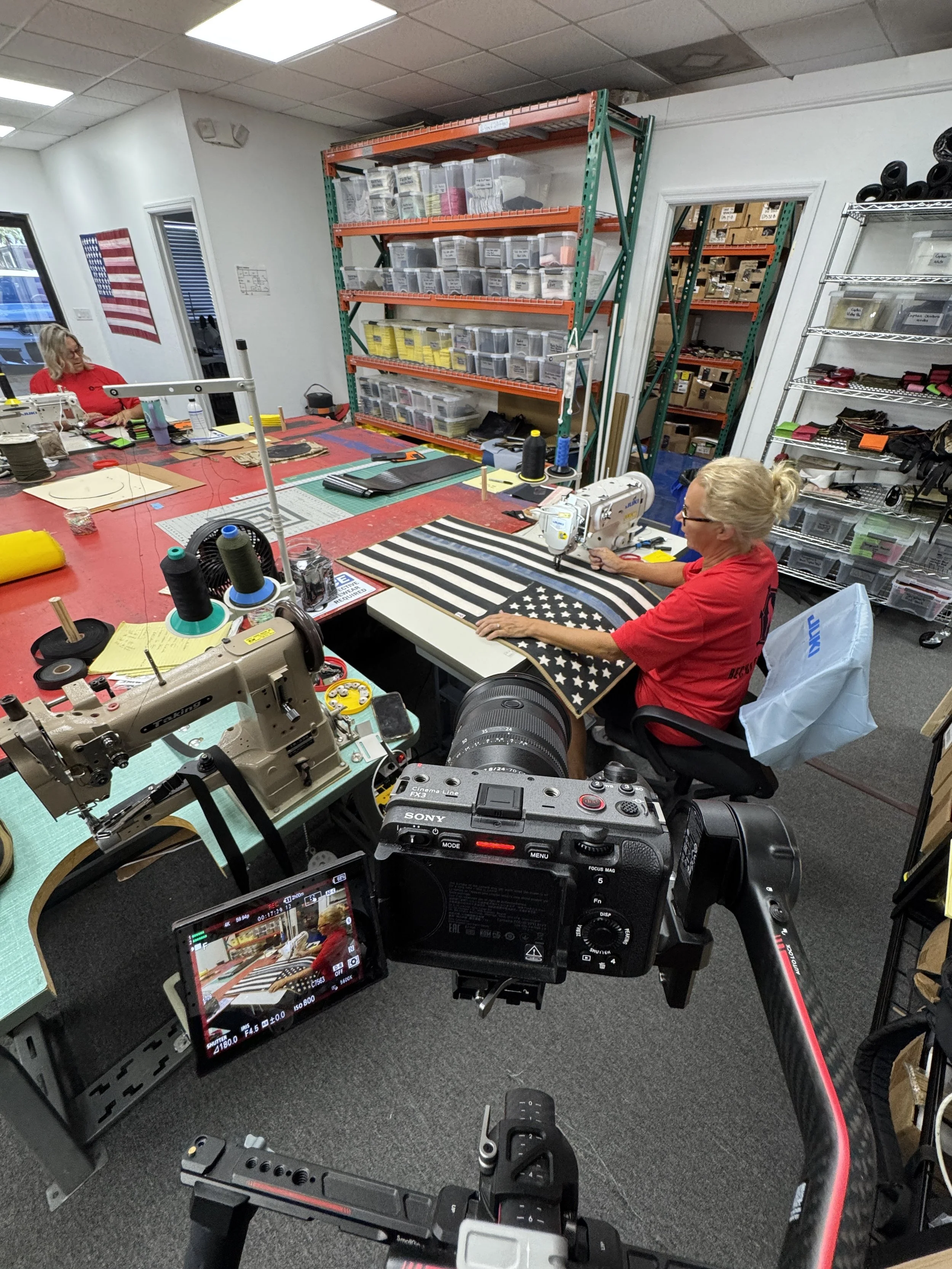 Two women working at a crafting or sewing workspace. One woman is seated at a sewing machine with American flag fabric, and the other woman is sitting nearby with a cutting mat and materials. The room has storage shelves filled with supplies and tool