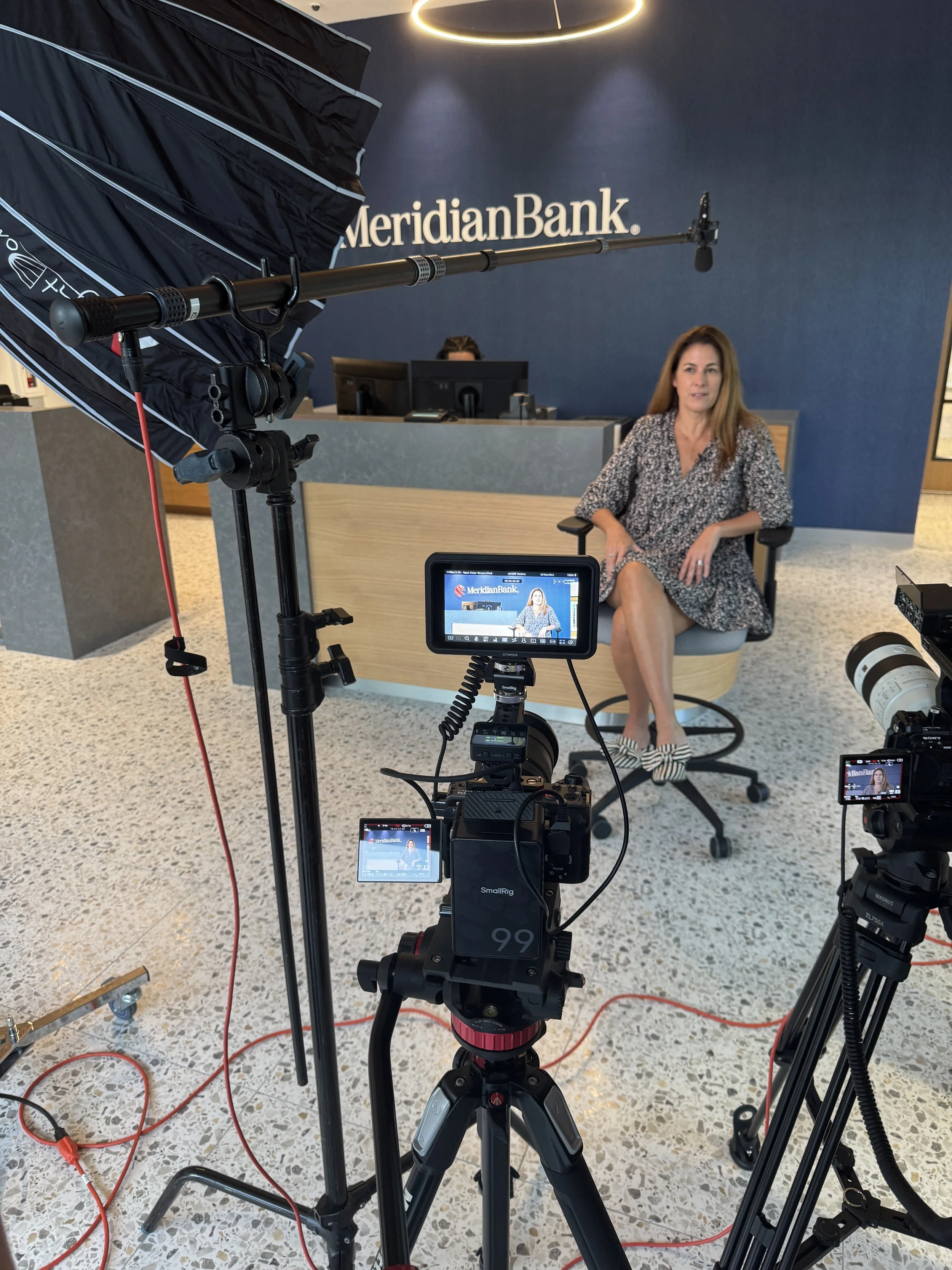 A woman sitting in a conference room with a blue wall behind her that has 'MeridianBank' written on it. There is professional video recording equipment, including cameras and lights, set up around her for a recording or interview.