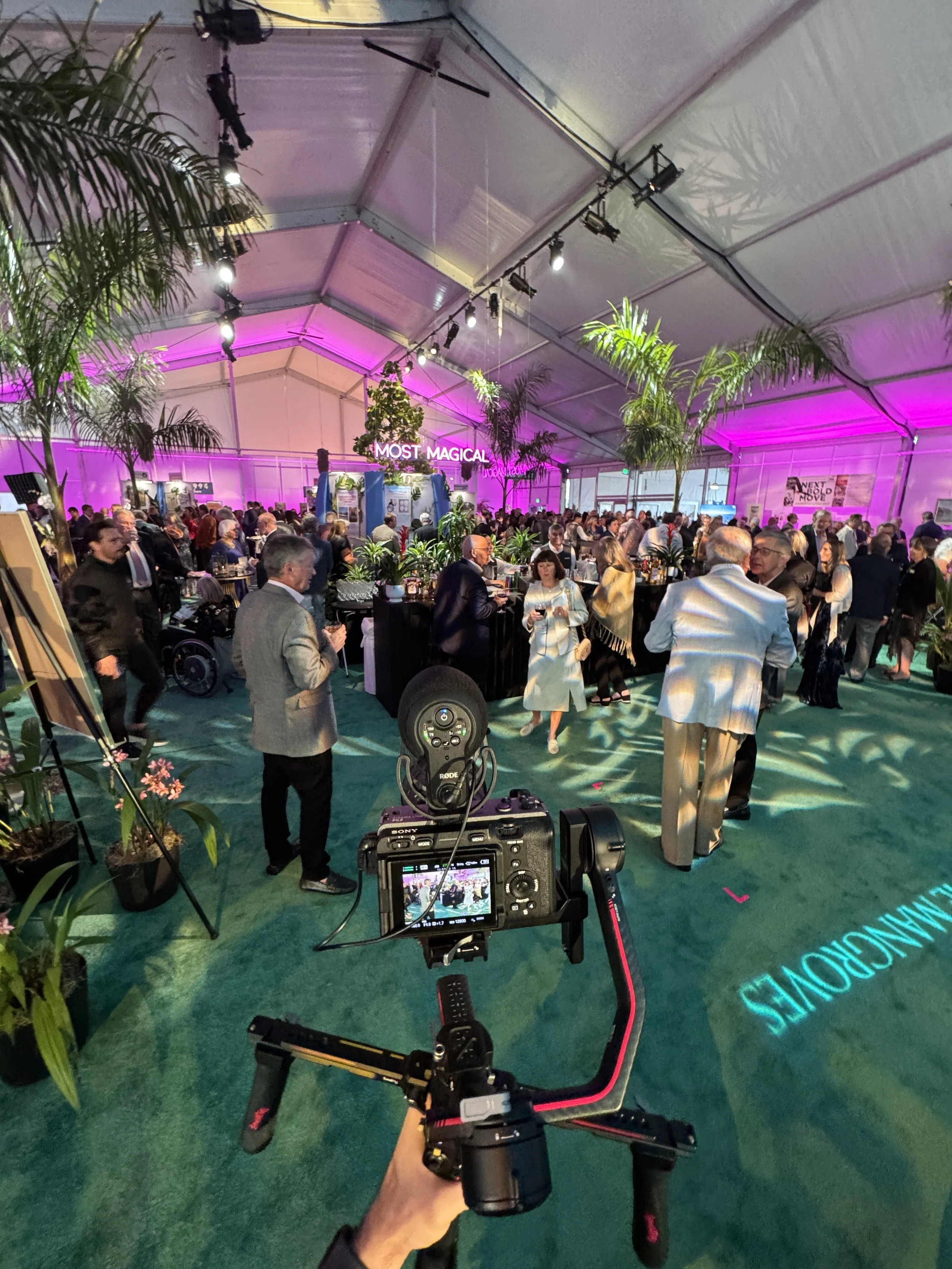 View from a camera mounted on a gimbal capturing a large indoor event with many people. The event space is decorated with plants and purple lighting, with a sign displaying "Most Magical" and various booths or displays in the background.
