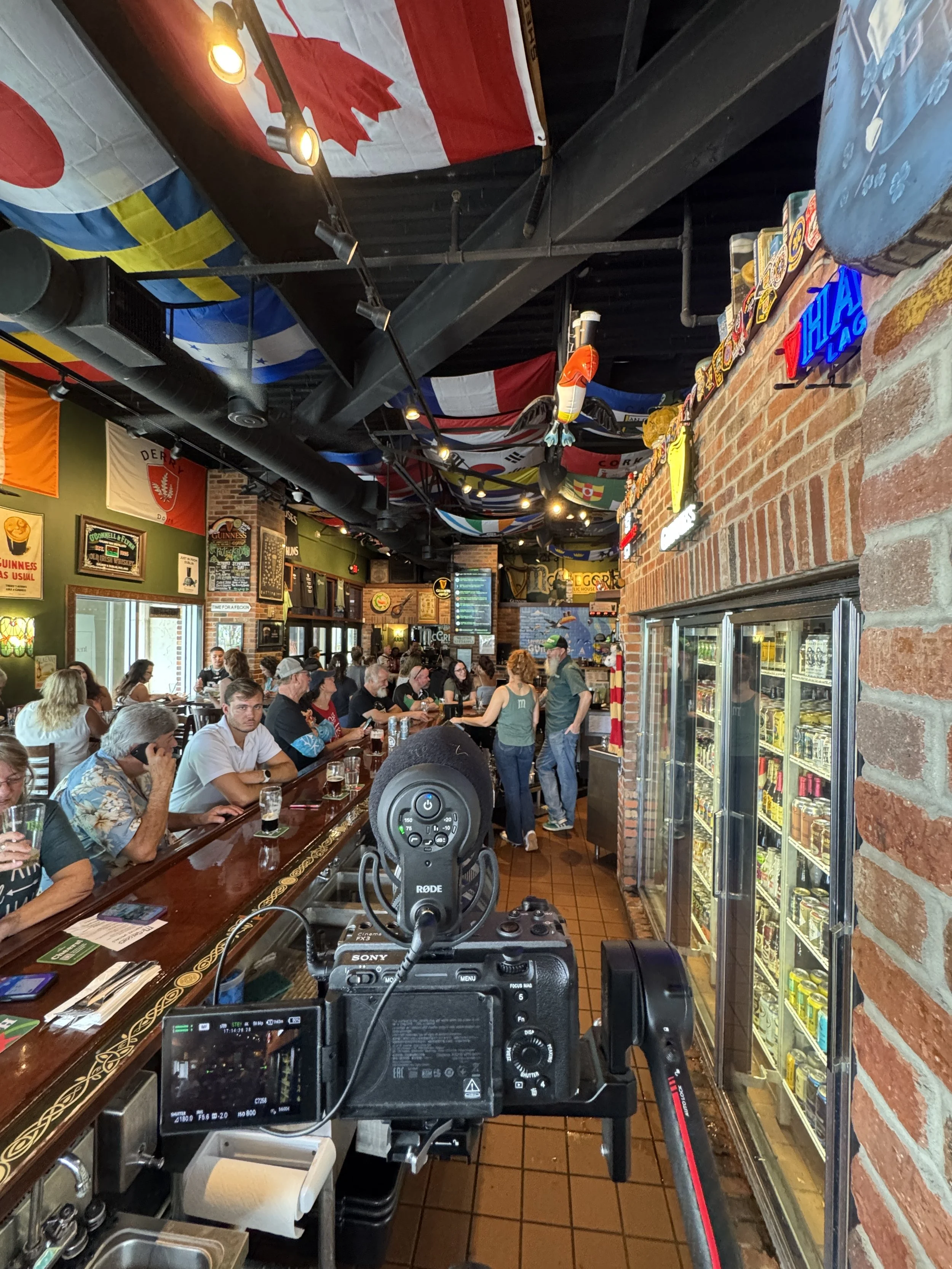 Interior of a busy bar or restaurant with patrons sitting at a wooden counter, flags hanging from the ceiling, and a camera setup in the foreground.