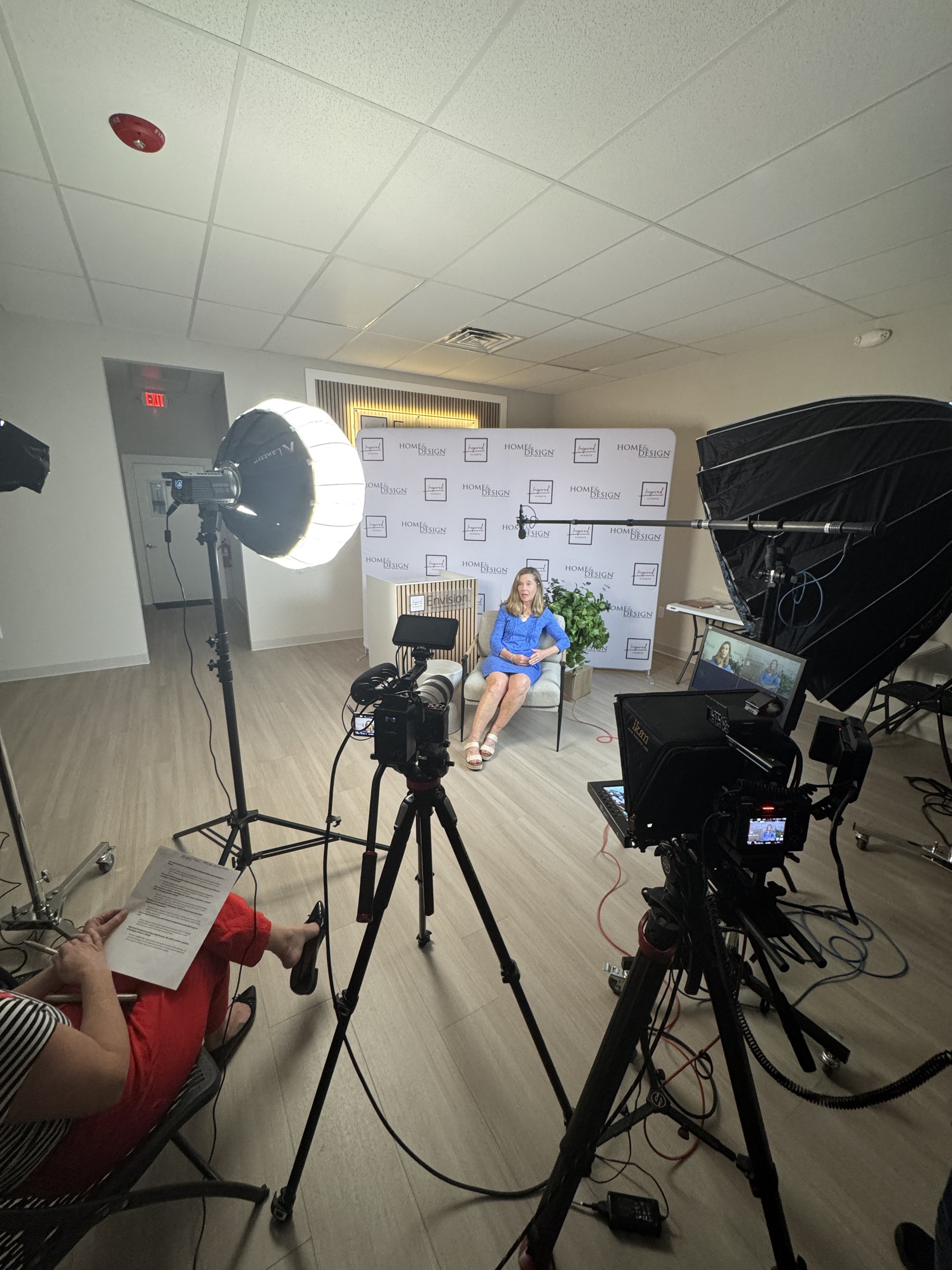 A woman in a blue dress seated on a chair and speaking in a studio setup with lighting equipment, a backdrop with patterned designs, and a person in red pants holding a paper.