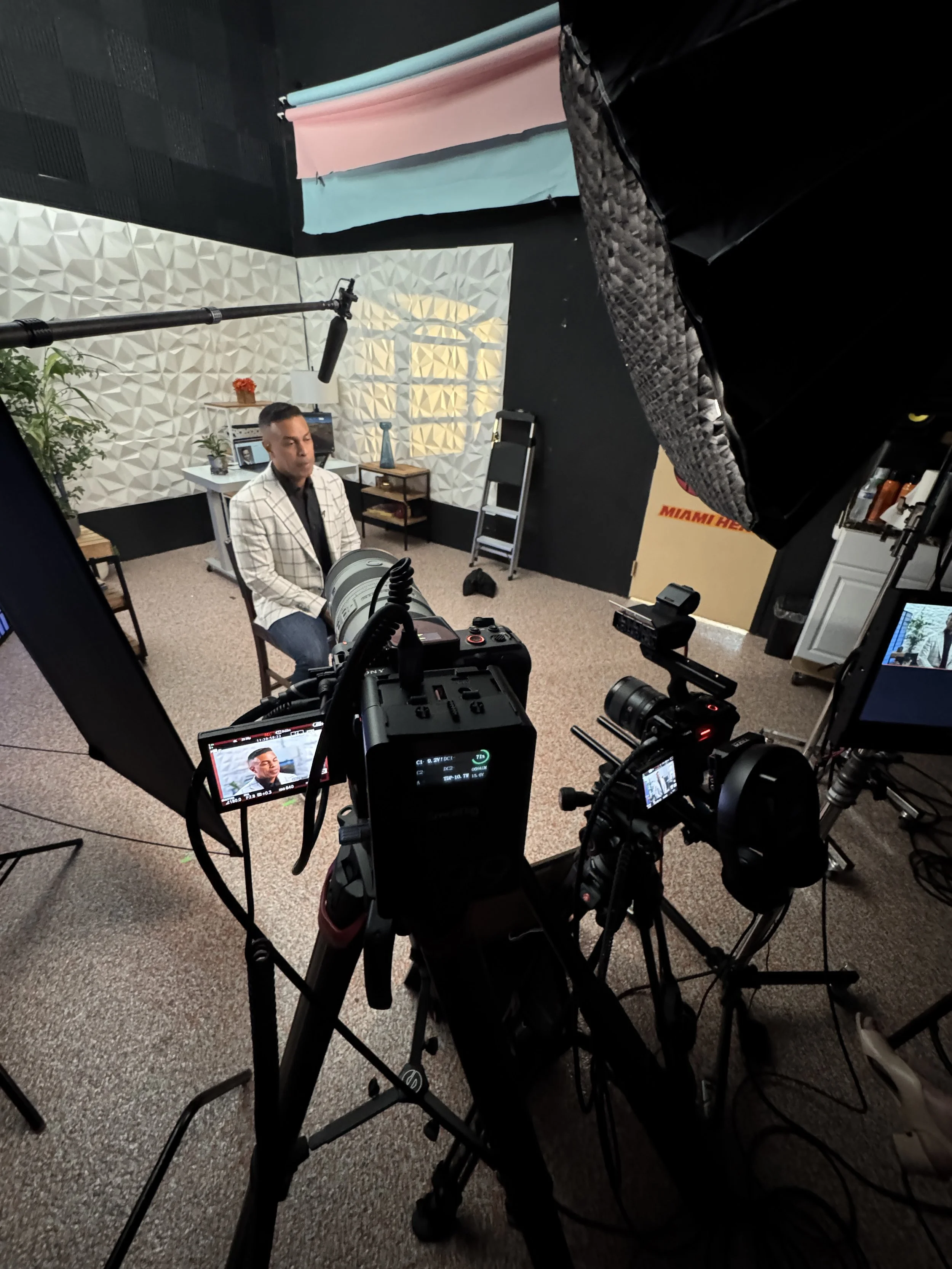A man sitting on a chair in a recording studio, surrounded by professional camera equipment, with a textured white wall and plants in the background.