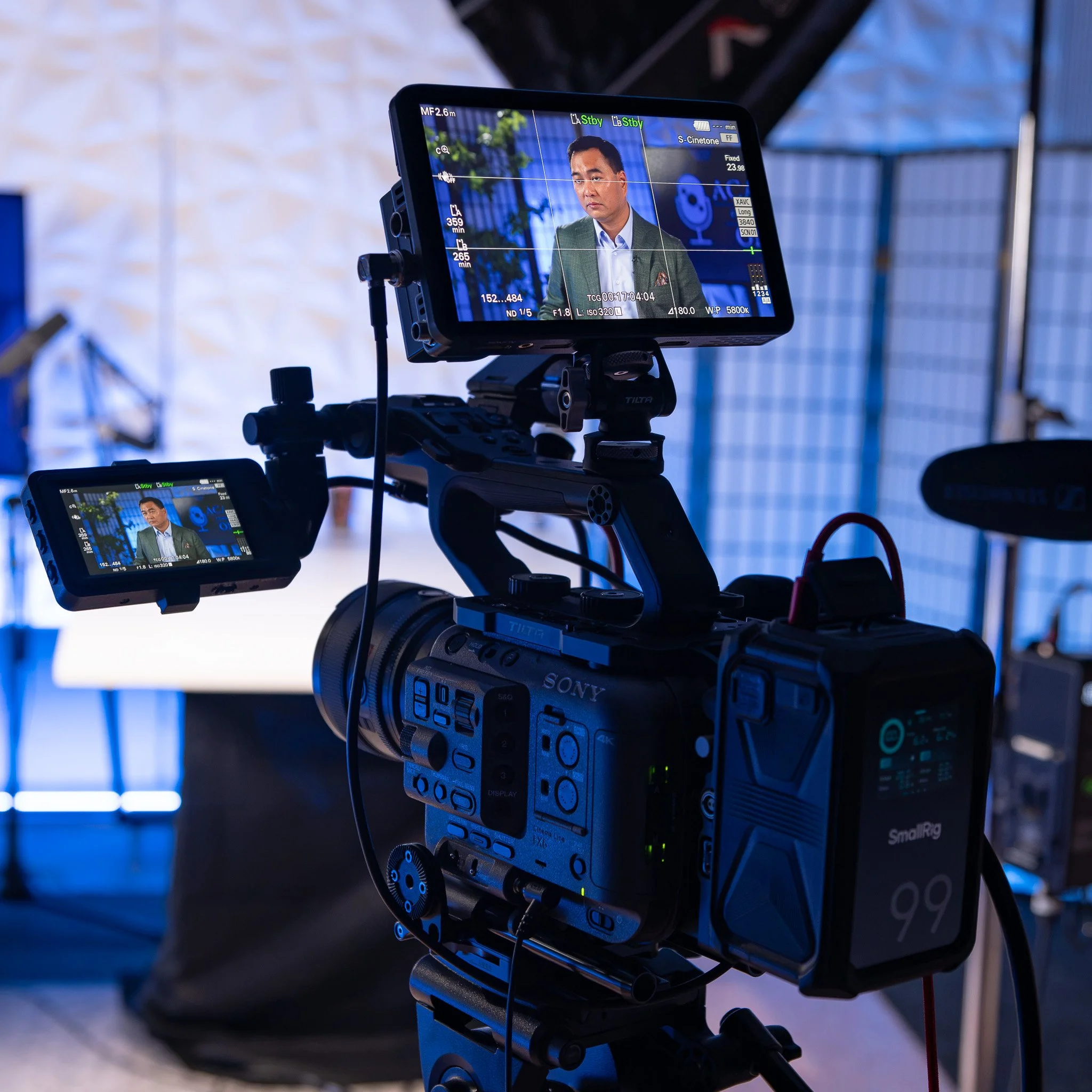 Close-up of a professional video camera monitor showing a man in a suit and tie, during what appears to be a video shoot or interview, with blue and green lighting in the background.