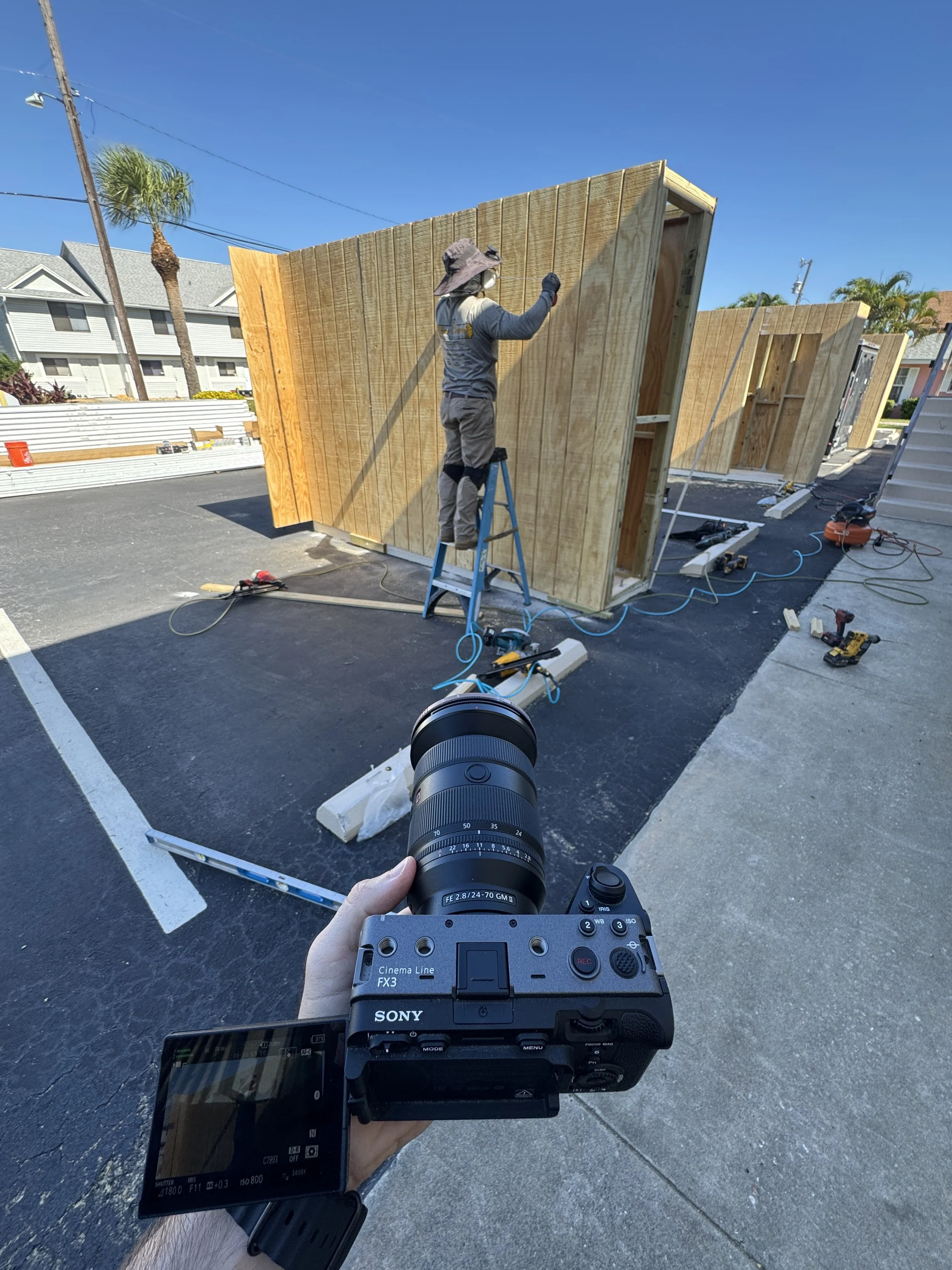 A construction worker standing on a ladder installing wooden walls on a small building in a parking lot, with tools and equipment scattered around.