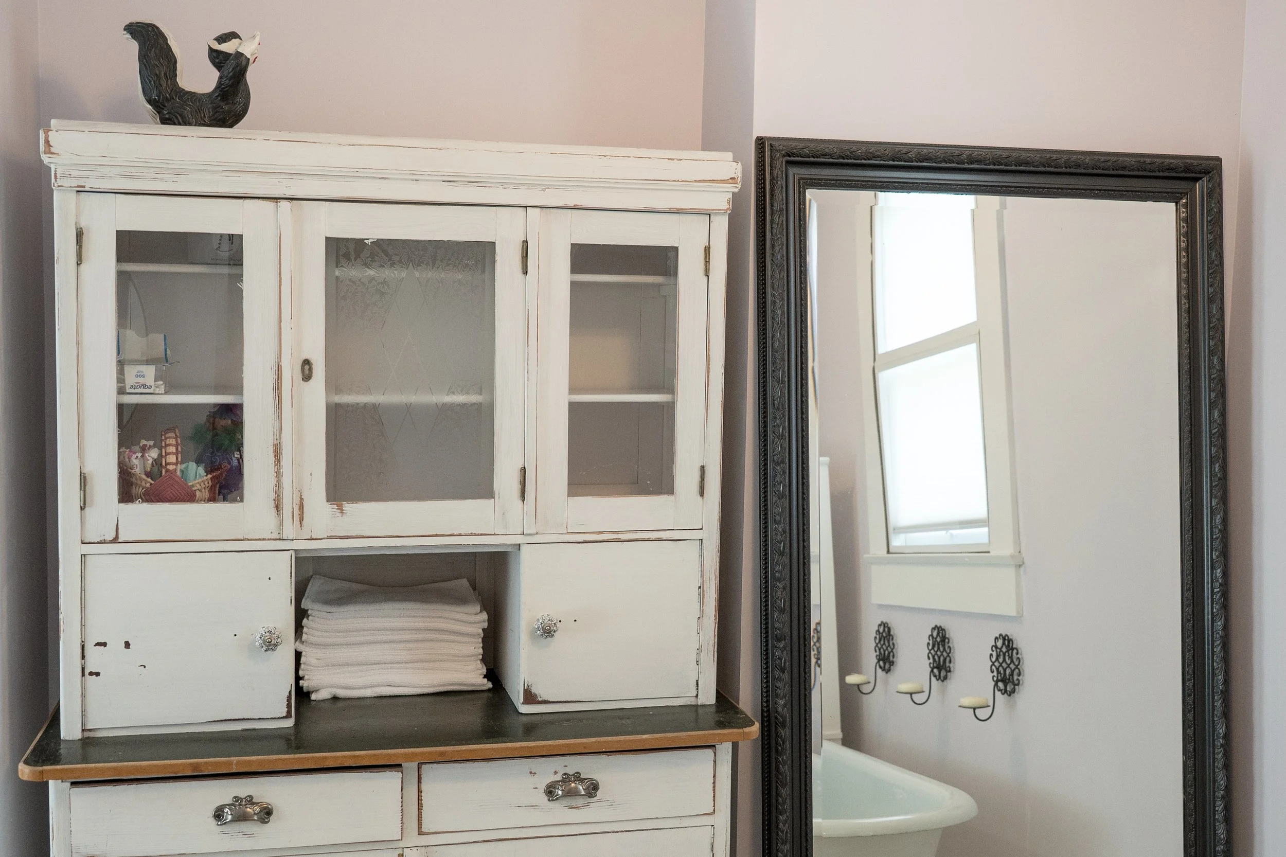 A vintage white glass cabinet with distressed paint, four doors, and decorative hardware, topped with a ceramic cat figurine. Next to it is a large, ornate mirror leaning against the wall, reflecting a window with blinds and a radiator.
