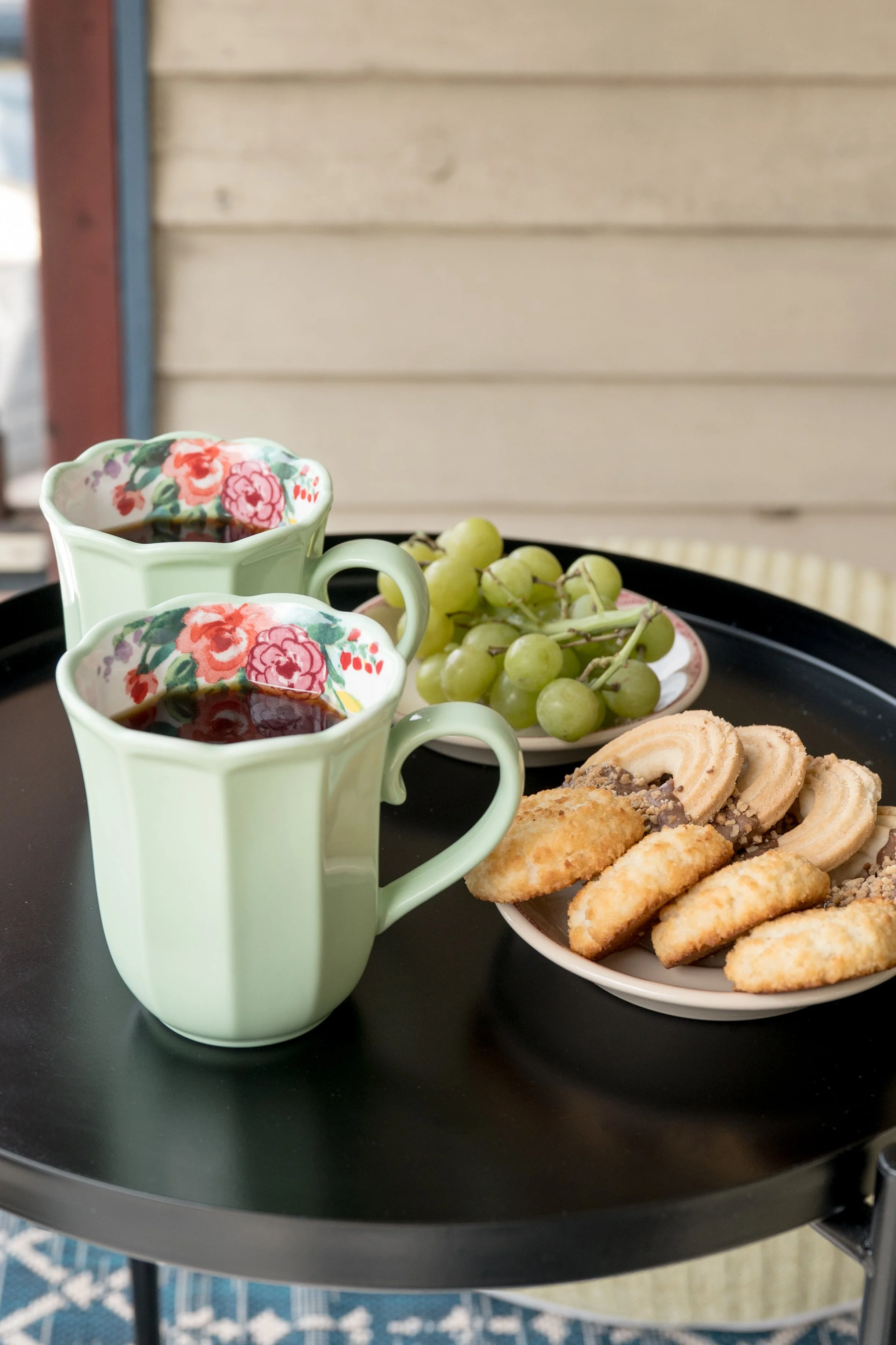 Two pale green floral teacups filled with dark tea, placed on a black tray alongside a plate of green grapes, cookies, and pastry sticks, against a wooden and outdoor background.