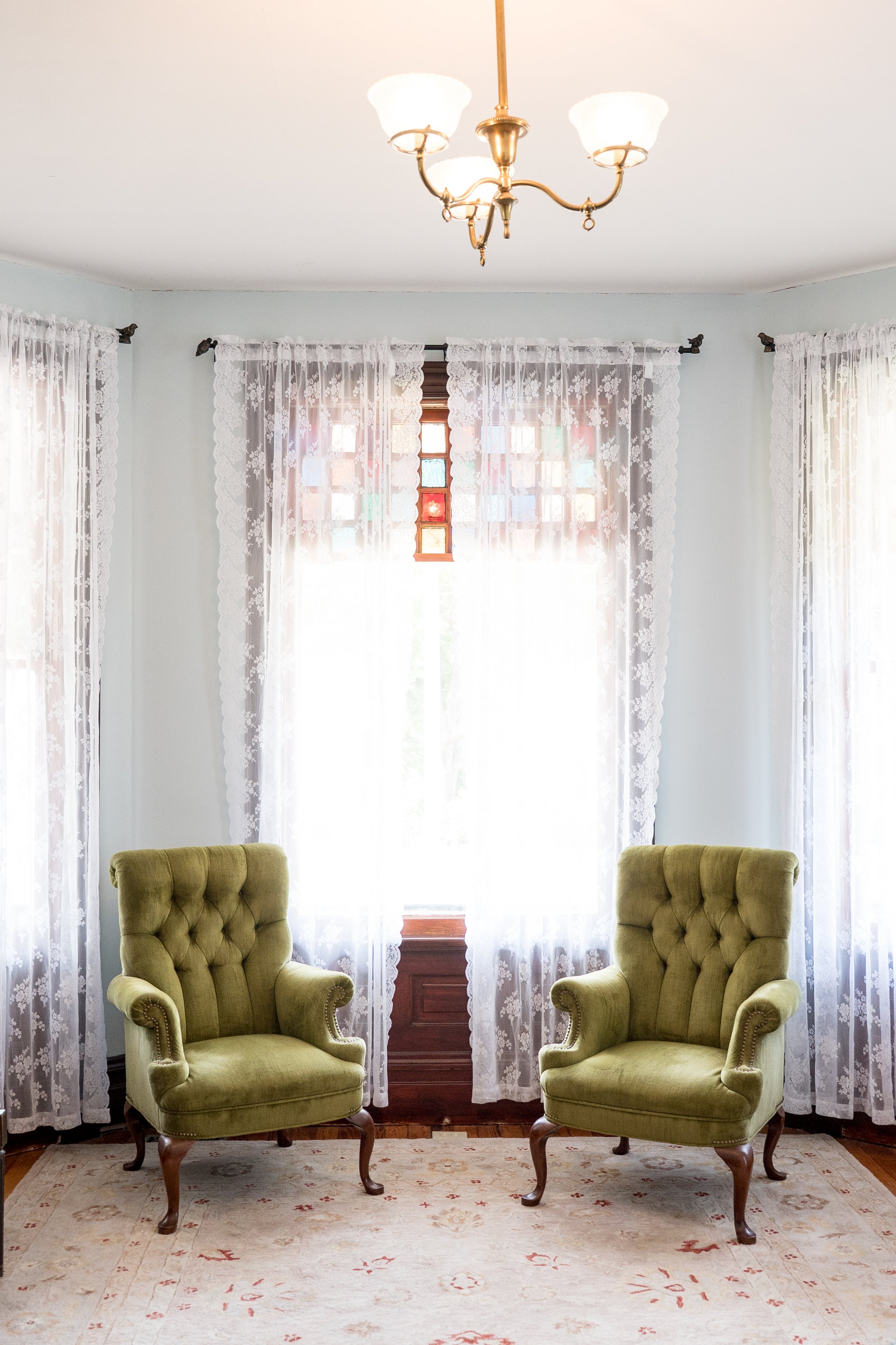 Living room with two green upholstered armchairs, lace curtains, a stained glass window, hardwood flooring, and a ceiling chandelier.