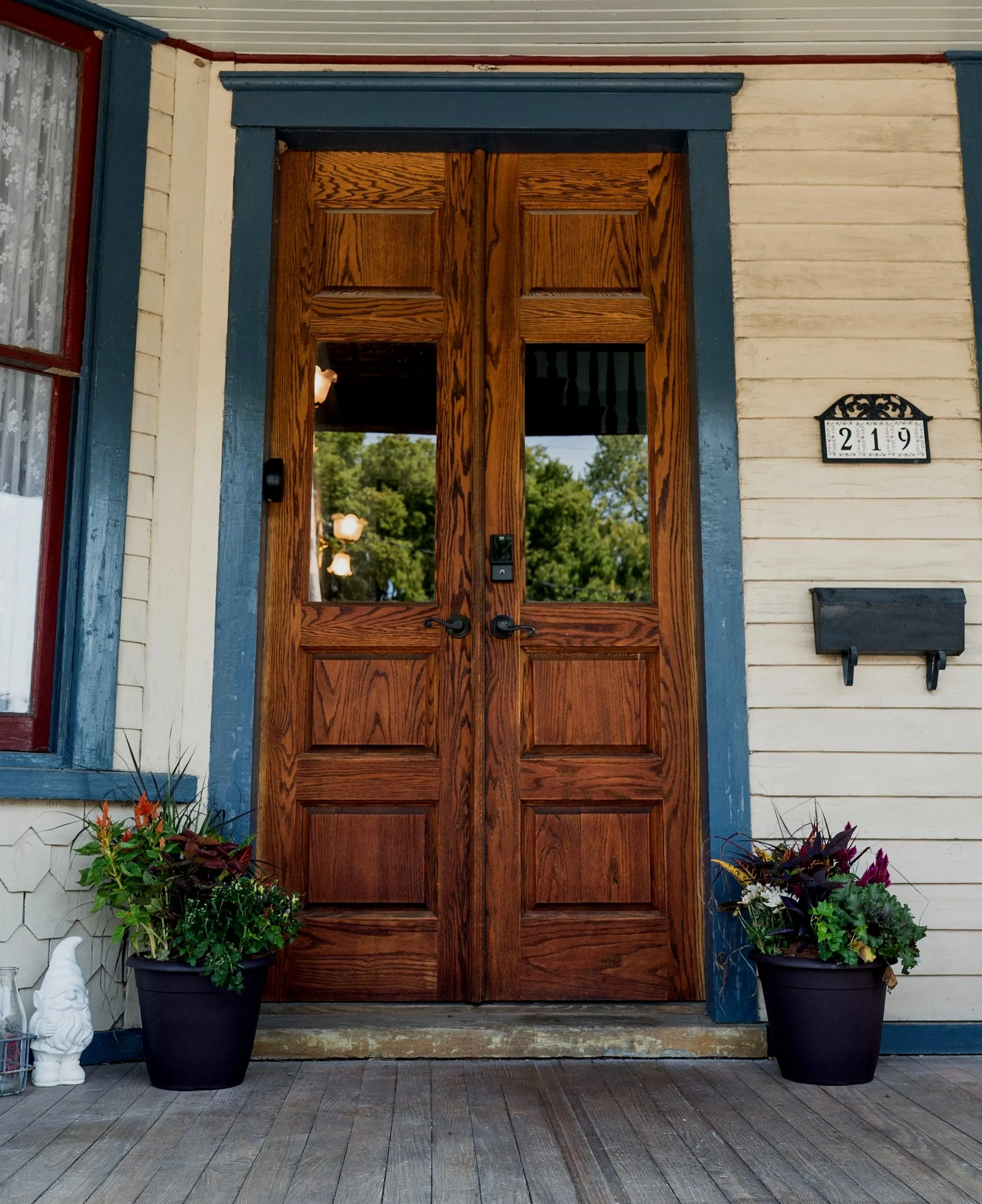 Wooden front door with glass panels, flanked by two potted plants on a porch, with house number 219 on the wall.