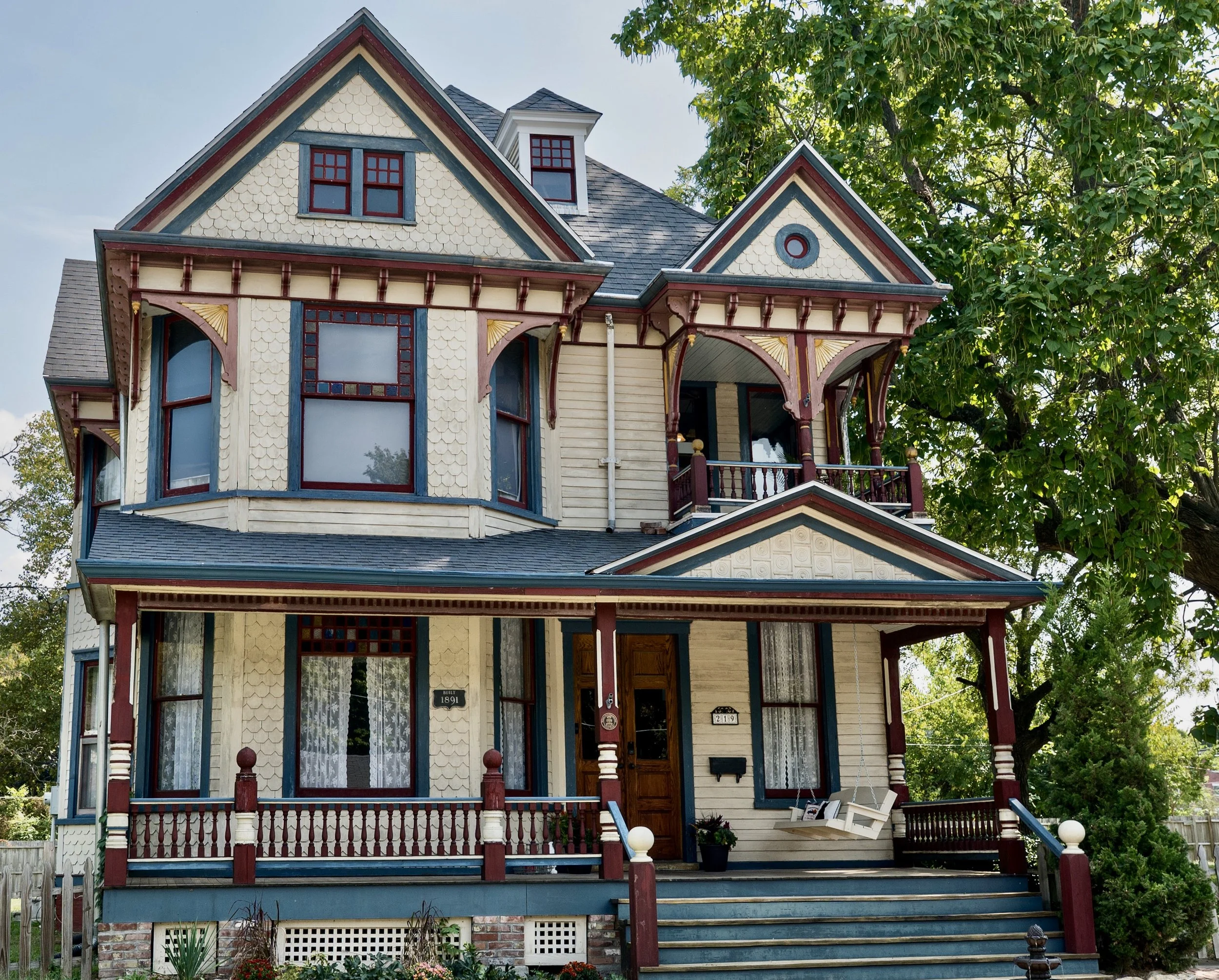 A Victorian-style house with ornate trim, multiple gables, bay windows, and a small porch. The house is painted cream with blue, red, and yellow accents. There are two signs on the front door with the numbers 1891 and 219, and a white bench is visible on the porch. The house is surrounded by mature trees and greenery.