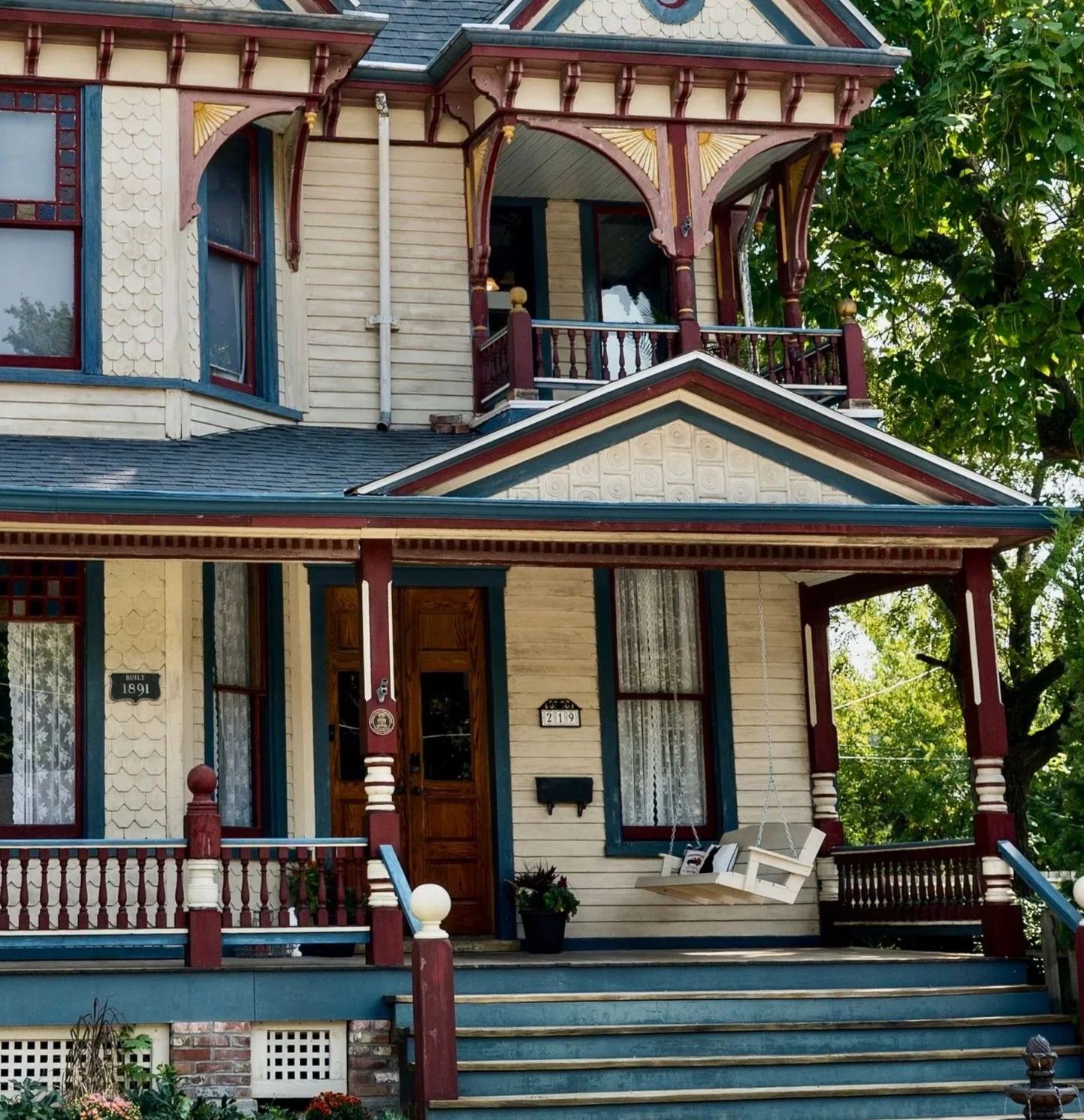 A vintage house with Victorian architecture, featuring a front porch with a swing, intricate woodwork, bay windows, and detailed trim painted in cream, red, and blue. The house has a small set of stairs leading to the entrance, with trees and greenery in the background.