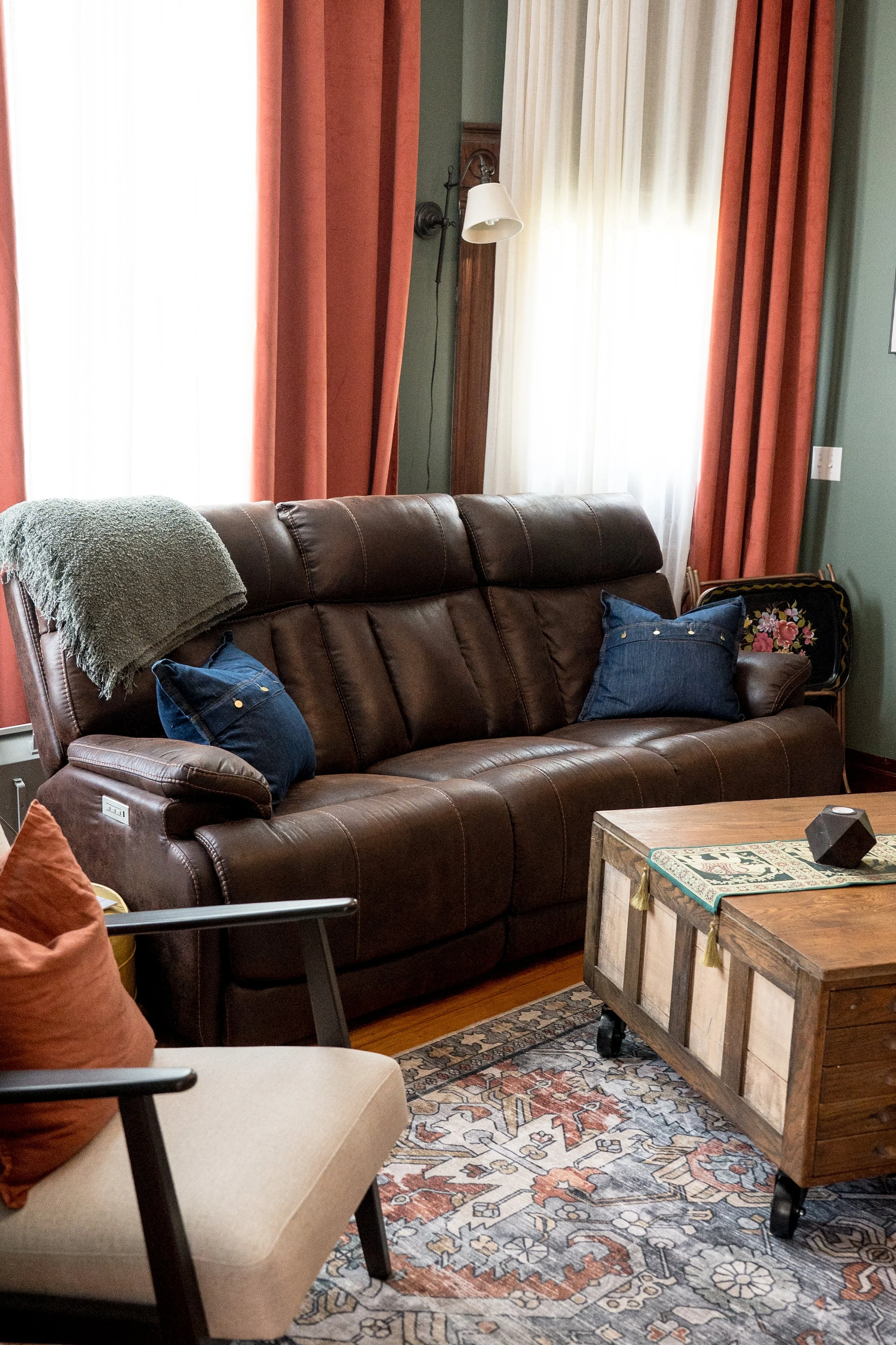 Living room with a brown leather sofa, blue pillows, and a beige chair with a brown pillow. A wooden coffee table with a decorative table runner and a black speaker. Bright windows with red and white curtains. Extracted from a cozy, rustic-themed living space.