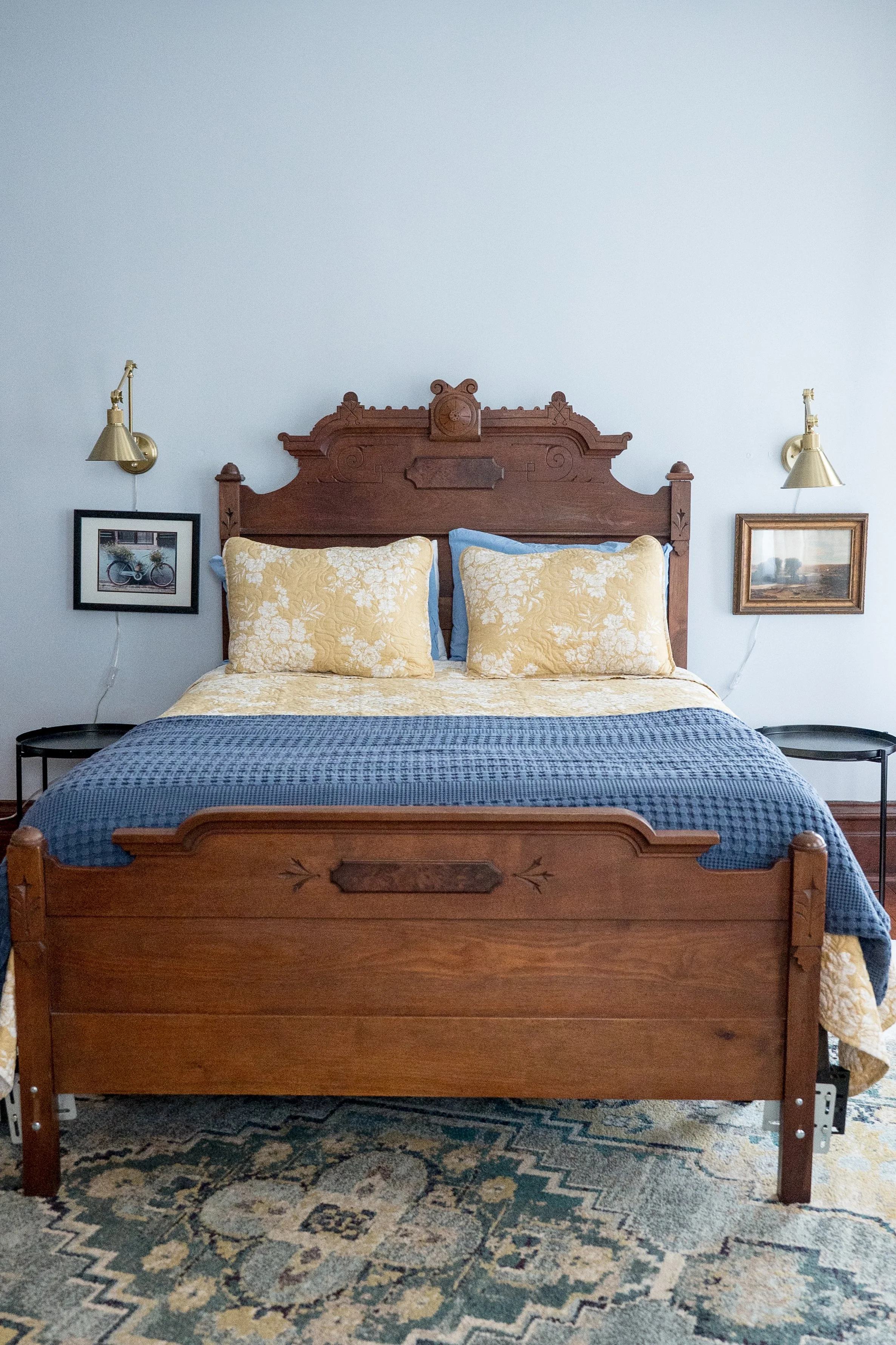 A wooden bed with a carved headboard features yellow floral pillows, a yellow floral bedspread, and a blue quilt. Two framed pictures hang on the wall, and two gold wall-mounted lamps illuminate the bed. A patterned rug covers the floor.