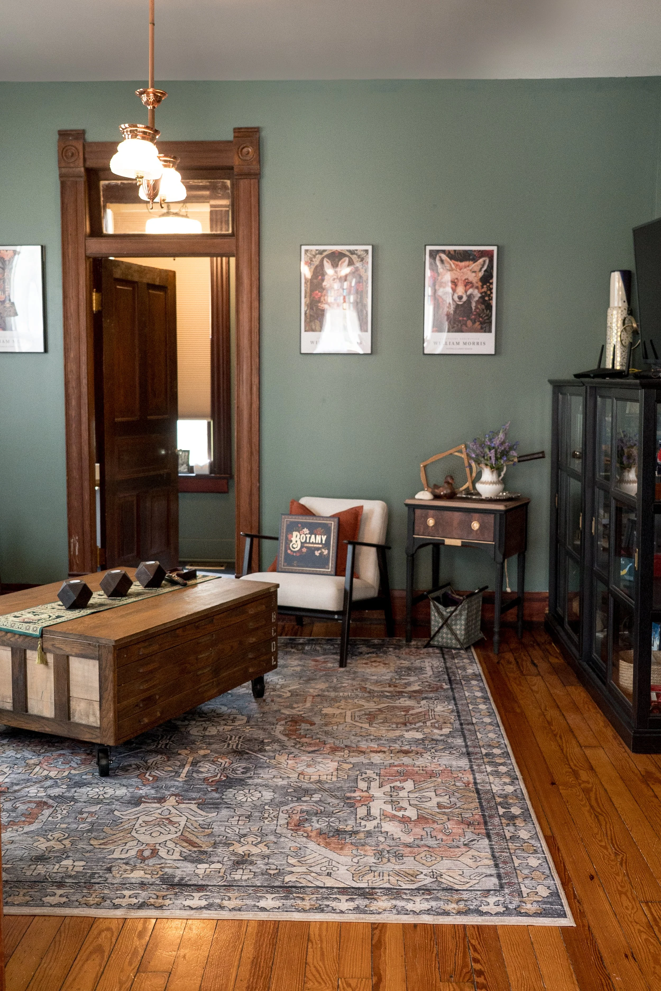 A cozy living room with a green wall, wooden trim, and vintage-style decor. There is a wooden coffee table with geometric black decorative objects, a beige armchair with an orange pillow, a side table with a vase of purple flowers, and framed animal art on the wall. A black cabinet with decorative items and a television is visible on the right. The room has hardwood flooring and a patterned area rug.