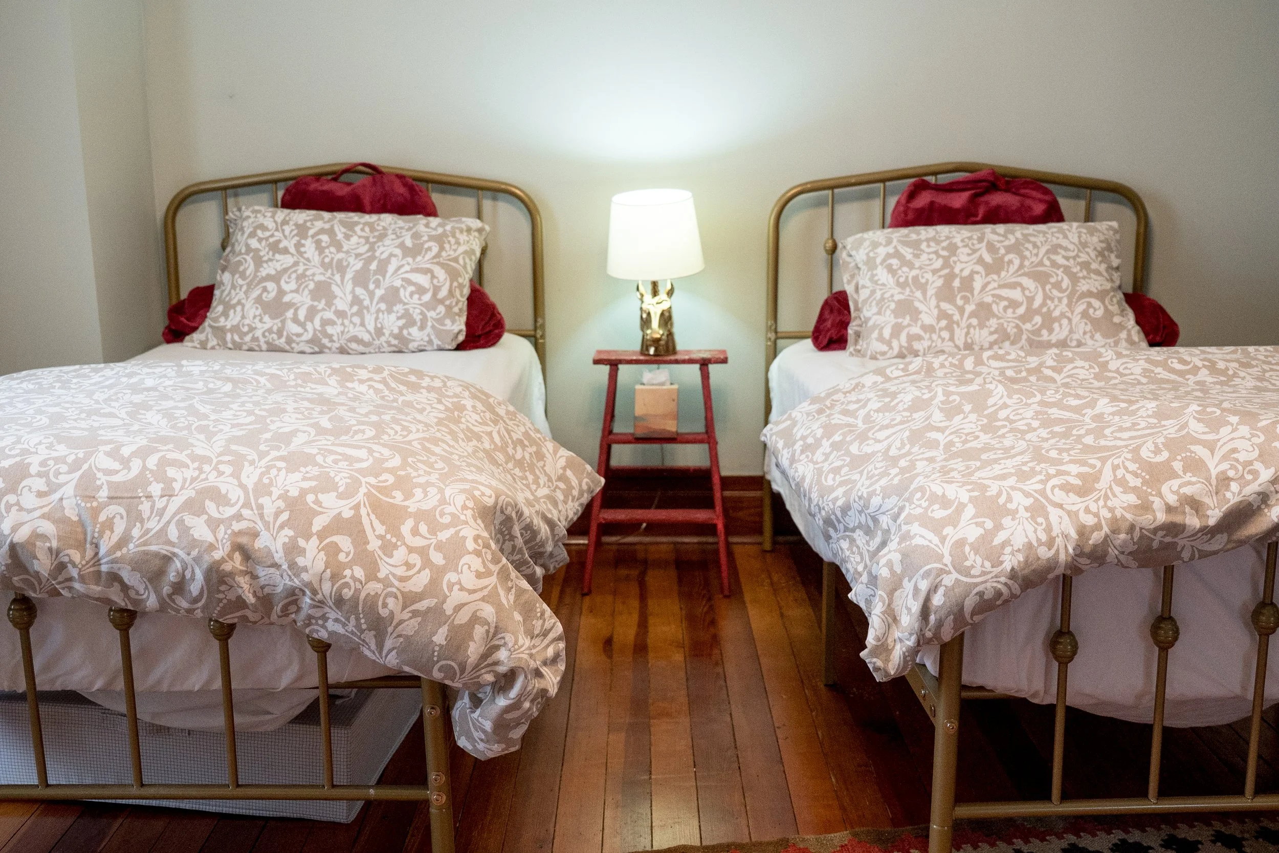 Two beds with beige and white patterned bedspreads and pillows, a nightstand with a lamp, and two red plush bags behind the pillows in a bedroom with wooden flooring.