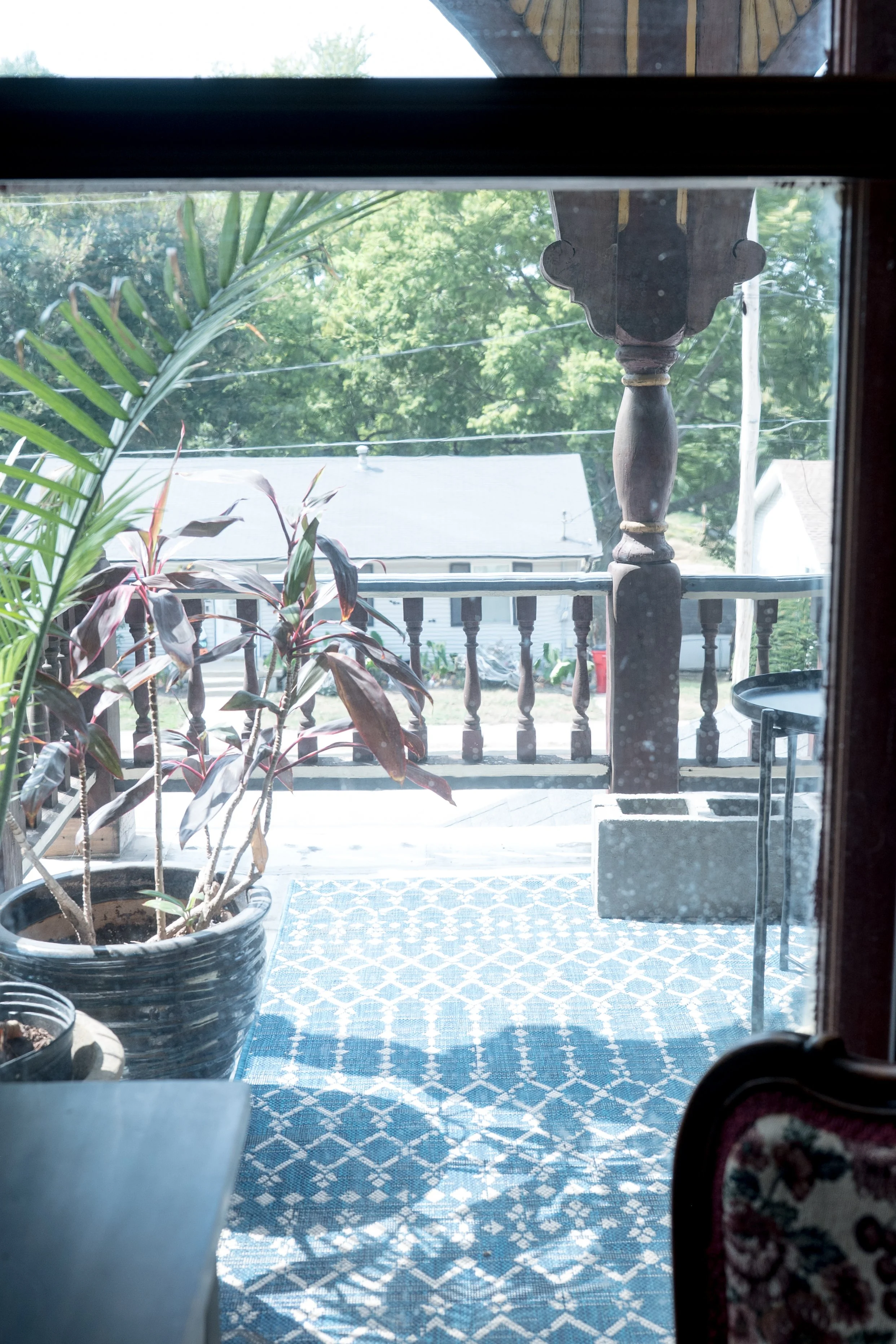 View from inside a house looking out through a glass door leading to a porch with plants, a wooden railing, and a neighboring house with a gray roof, surrounded by greenery.