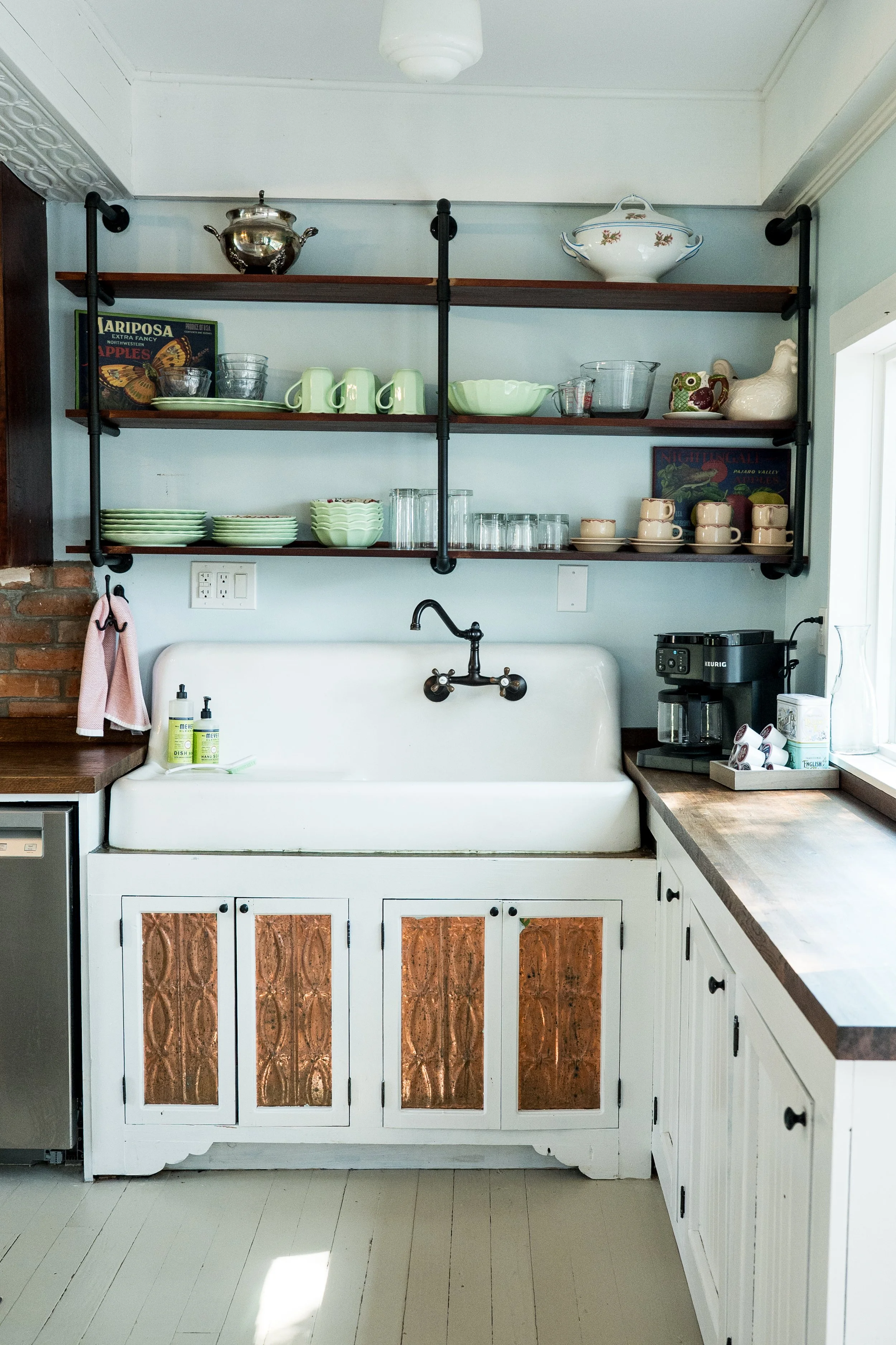Kitchen with open shelves holding dishes, cups, and decorative items, white cabinets with copper-colored panels, and a farmhouse sink with a black faucet.