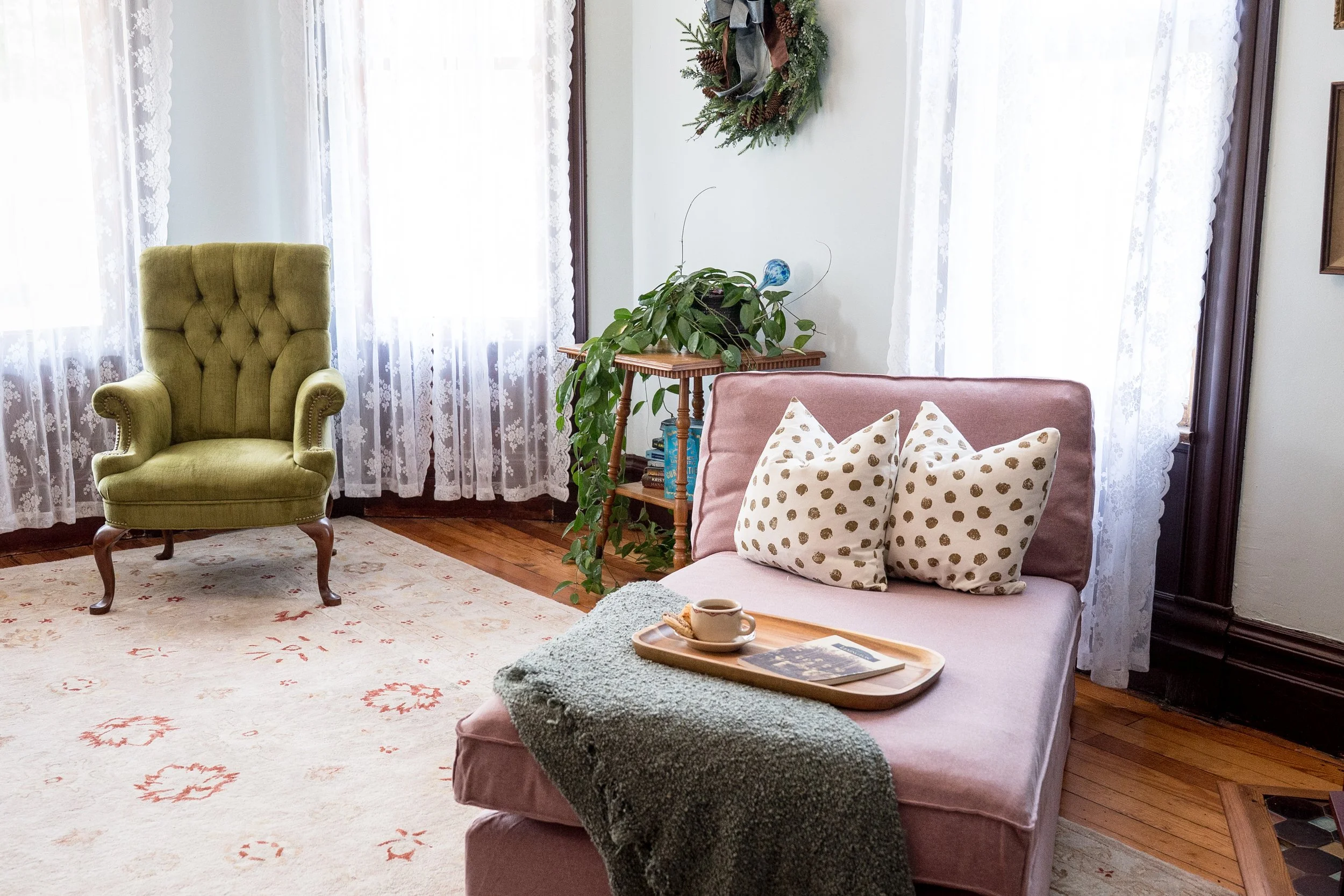 A cozy sitting room with lace curtains, a green velvet armchair, a pink chair with polka dot pillows, a small table with a cup, a tray, and a magazine, houseplants on a small table, and a holiday wreath on the wall.