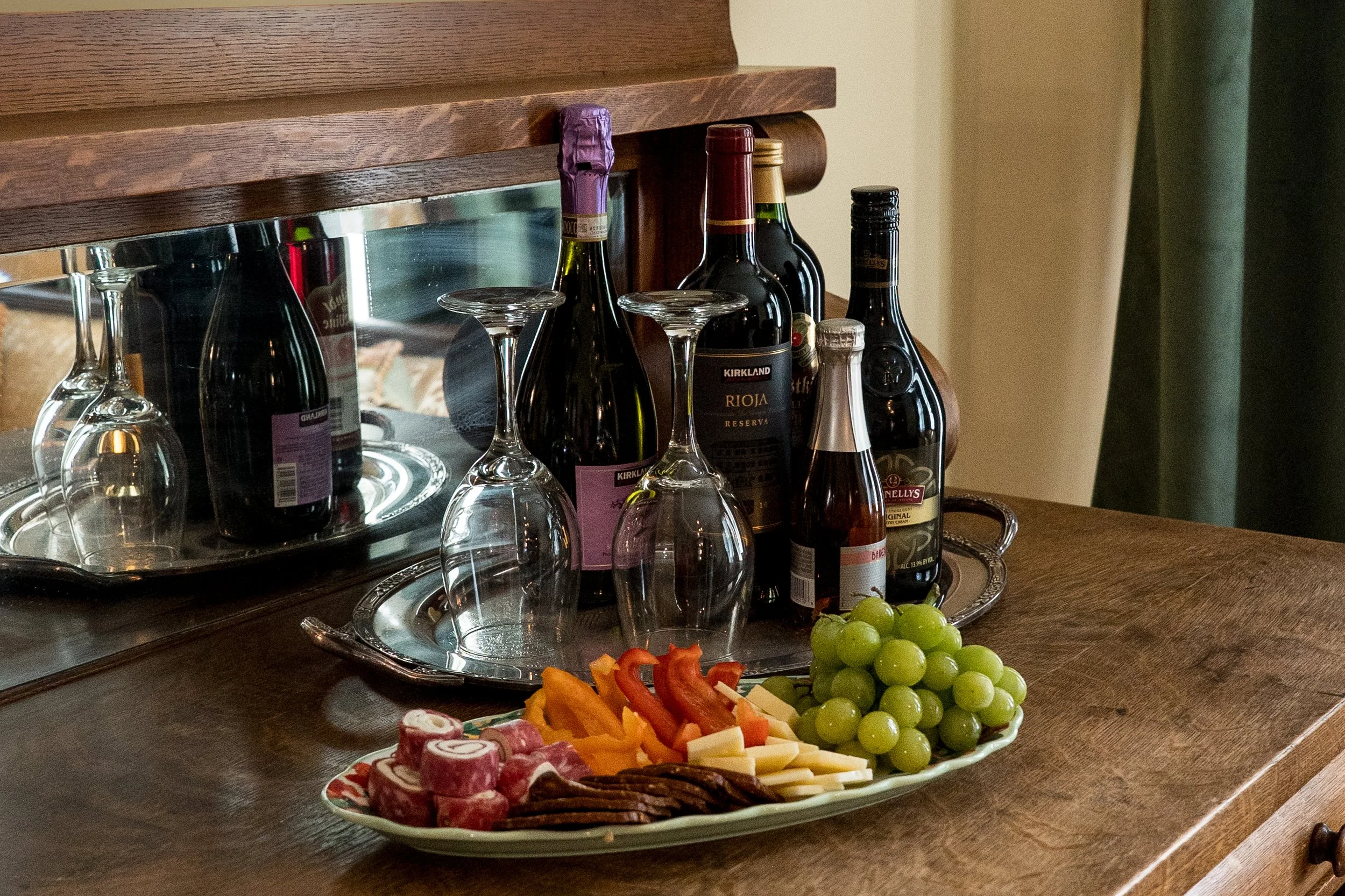 A wooden sideboard with a mirror decor and cold beverages in bottles, and a silver tray of green grapes and assorted sliced fruits and cheeses on a table.