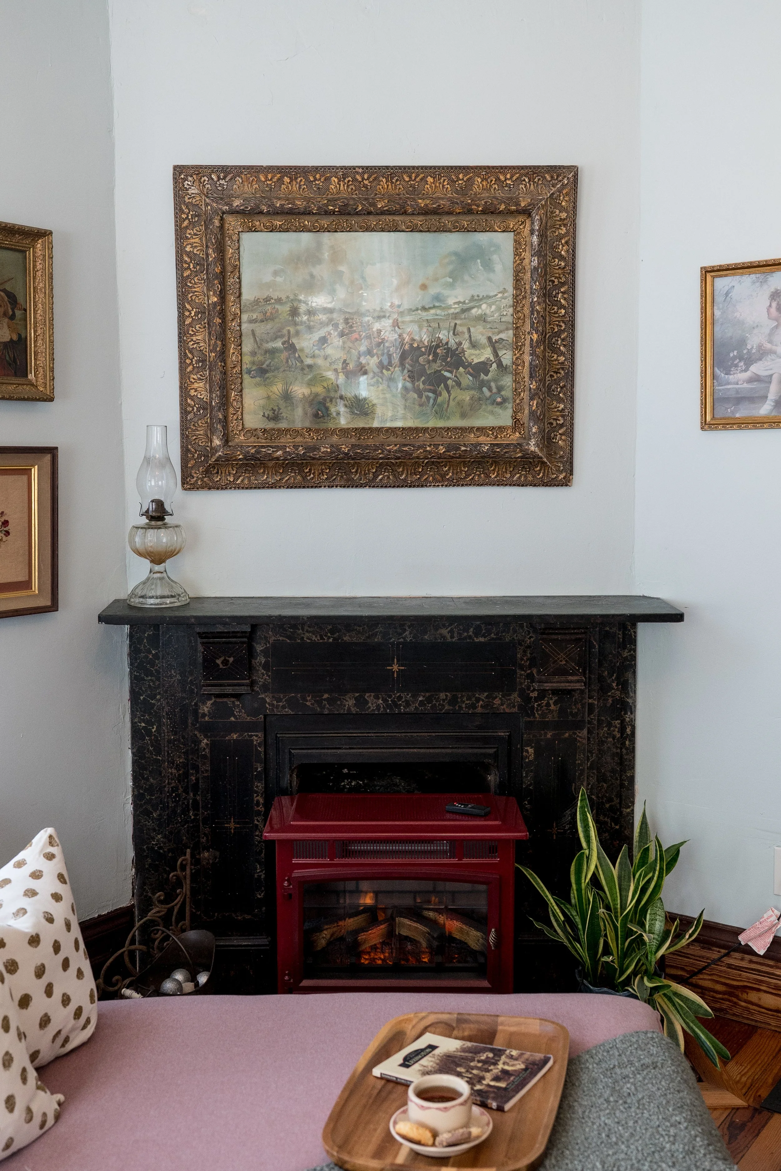 A cozy living room scene featuring a fireplace with a red heater insert, a large framed painting of a historical battle scene above the fireplace, a table with a mug and pieces of cookies, a gray upholstered piece of furniture, and a plant in a black pot in the corner.