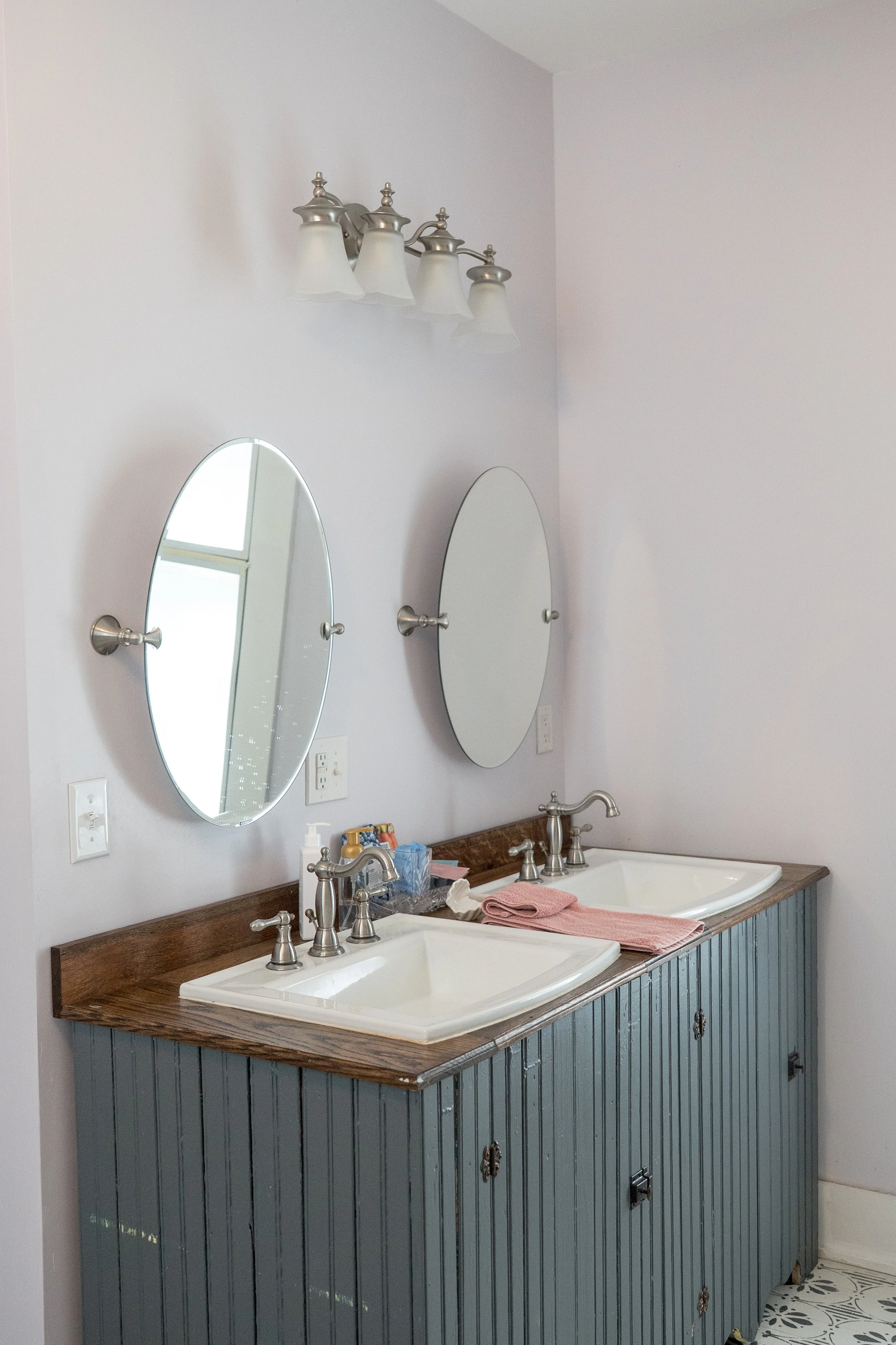 A bathroom vanity with two sinks, a wooden countertop, and blue beadboard cabinet doors. Two oval mirrors are mounted above the sinks, and soap dispensers, a pink towel, and toiletries are on the counter. White walls and a patterned tile floor are visible.