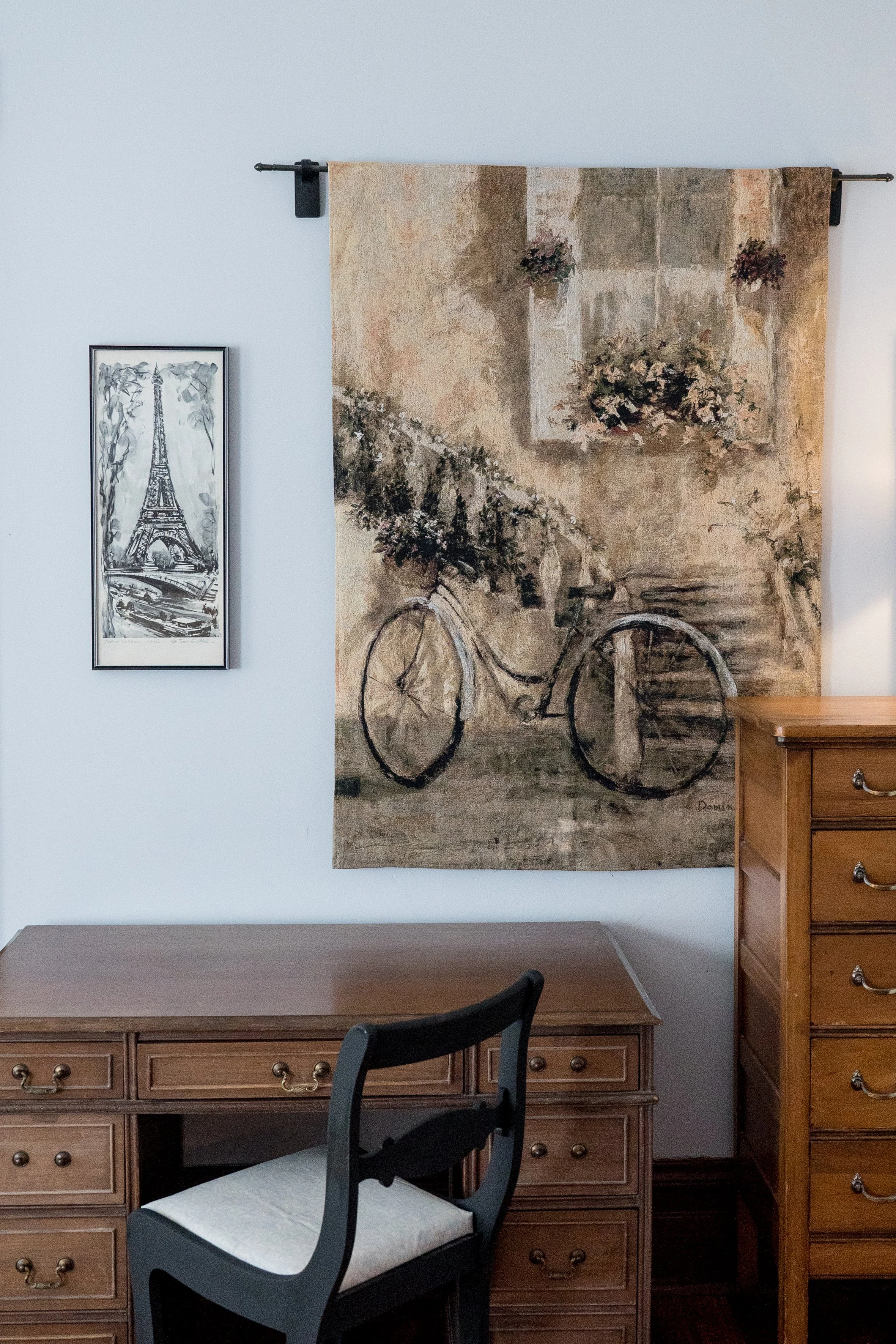 A room with a wooden desk and a black chair with a white cushion in front of it. A large wall hanging with a bicycle and flowers is above the desk. To the left, a framed black and white sketch of the Eiffel Tower is on the wall. There is a wooden dresser to the right of the wall hanging.