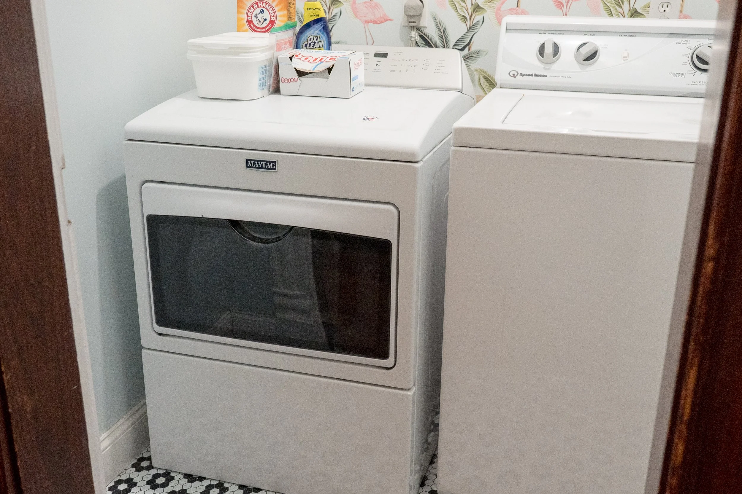 A laundry room with a Maytag front-loading washing machine beside a white top-loading dryer. On top of the washer, there is a white container, a box of Bounce dryer sheets, a pack of OxiClean, and some cleaning spray. The room has a floral wallpaper border and black and white hexagon tile flooring.
