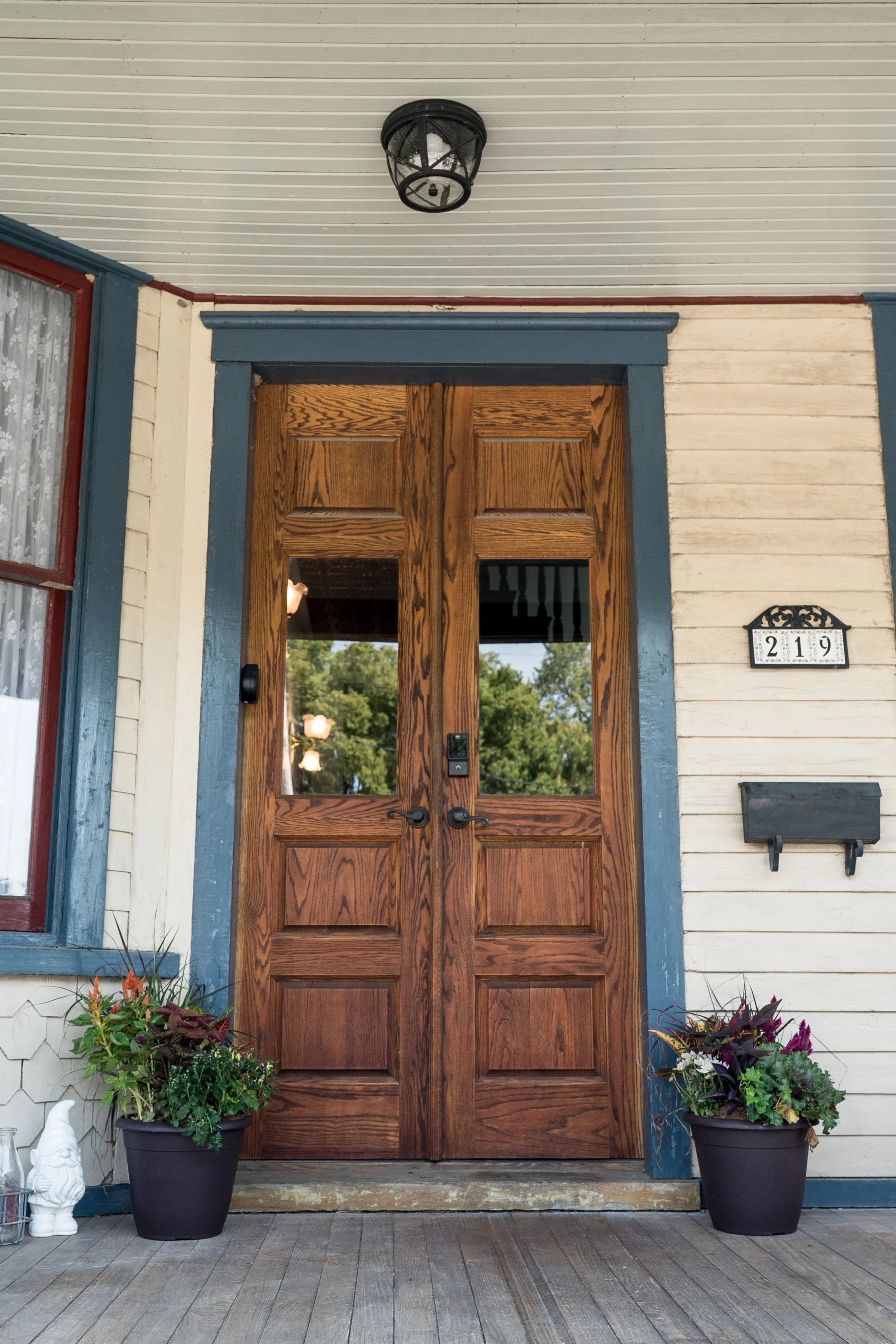 A front porch with a red door frame, double wooden doors, blue trim, and decorative house numbers 219. Potted plants with colorful flowers are on either side of the door, and a garden gnome statue is near the left plant. The porch has a wooden deck floor and a white ceiling with a black outdoor light fixture.