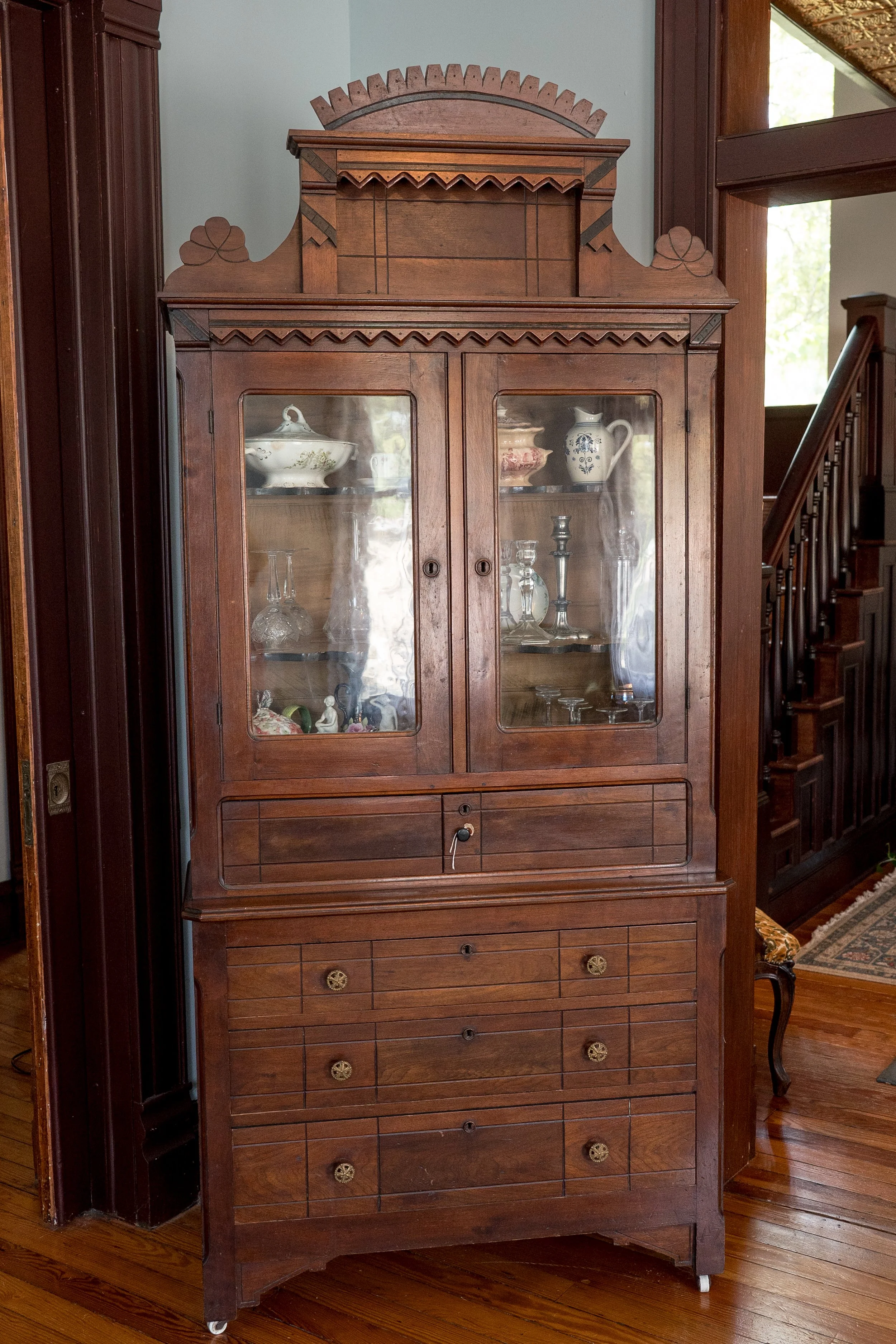 An antique wooden cabinet with glass doors displaying china and glassware, positioned against a light-colored wall in a room with hardwood floors.