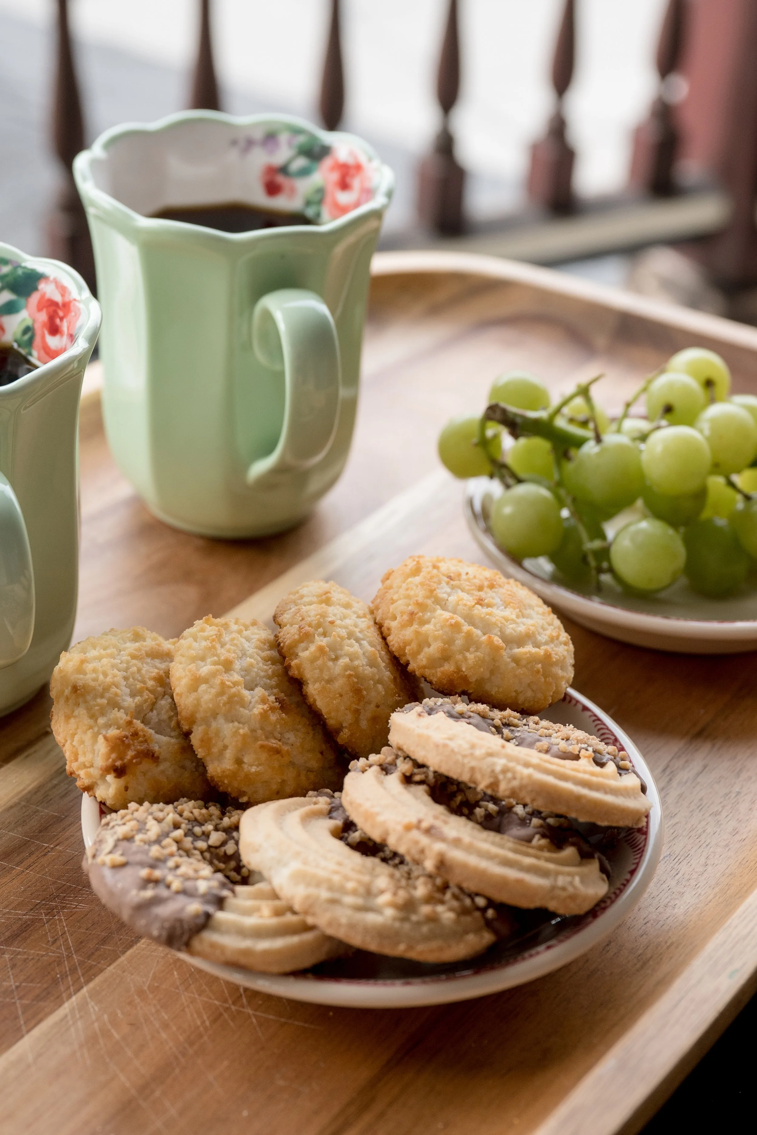 Plate of assorted cookies next to a bunch of green grapes, with two coffee mugs filled with coffee in the background.