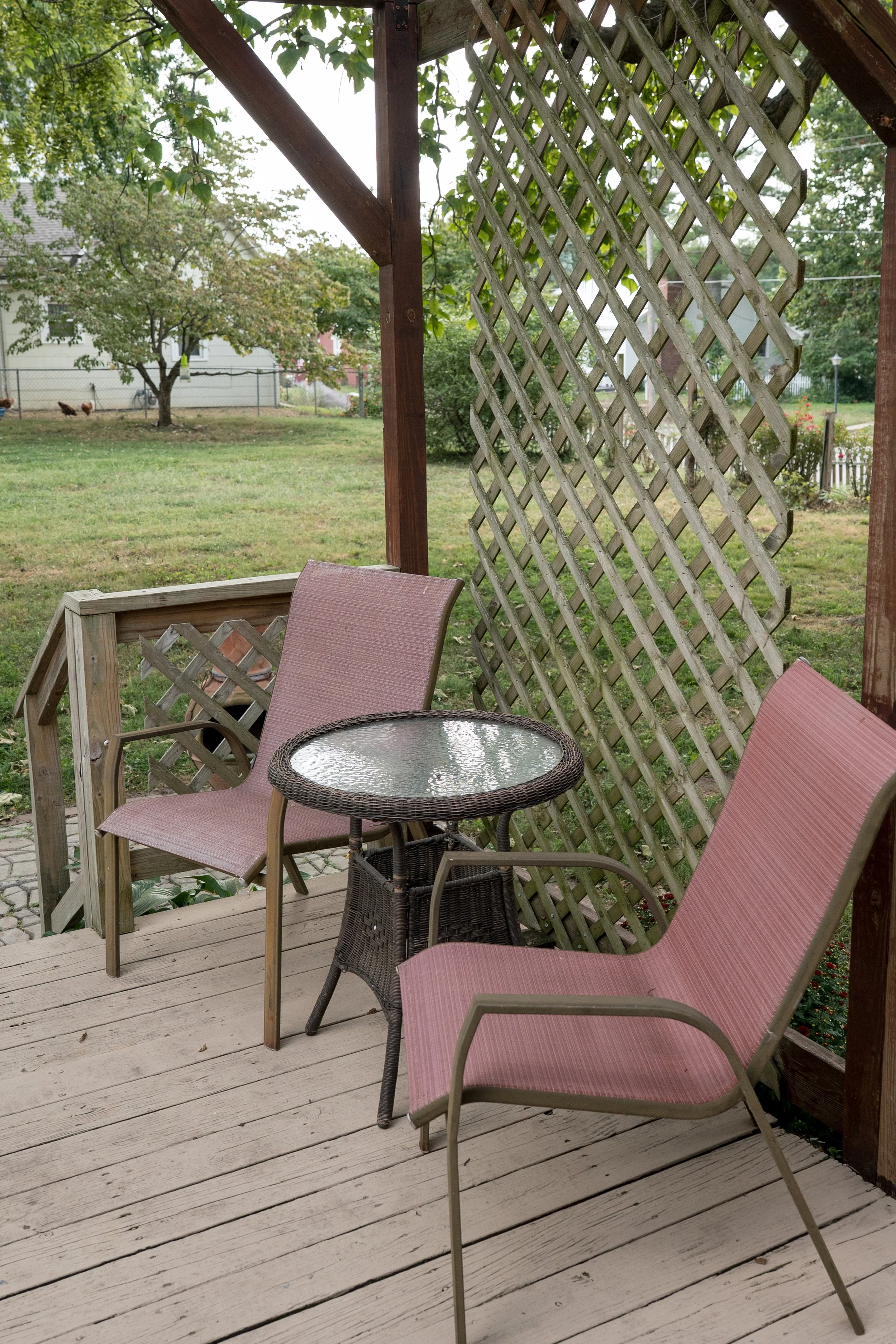 View of a backyard porch with pink outdoor chairs around a small round glass-top table, wooden railing, lattice privacy screen, and grassy yard with trees and chicken coop in the background.