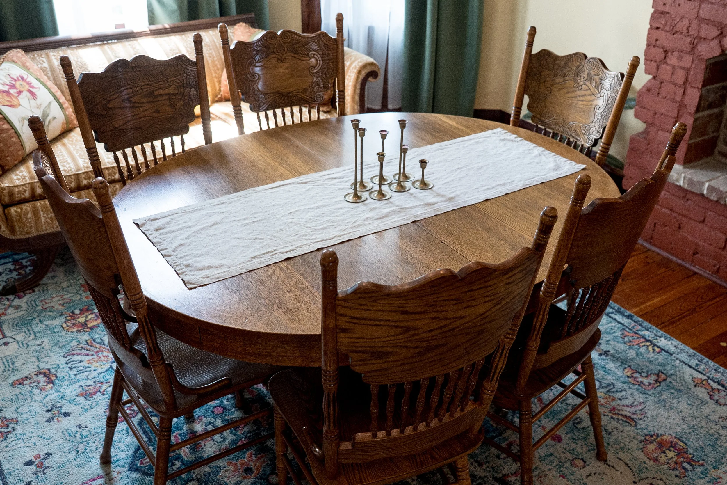 A wooden dining table with a white table runner and a candle holder centerpiece, surrounded by wooden chairs. In the background, there is a vintage style sofa with floral pillows and green curtains.