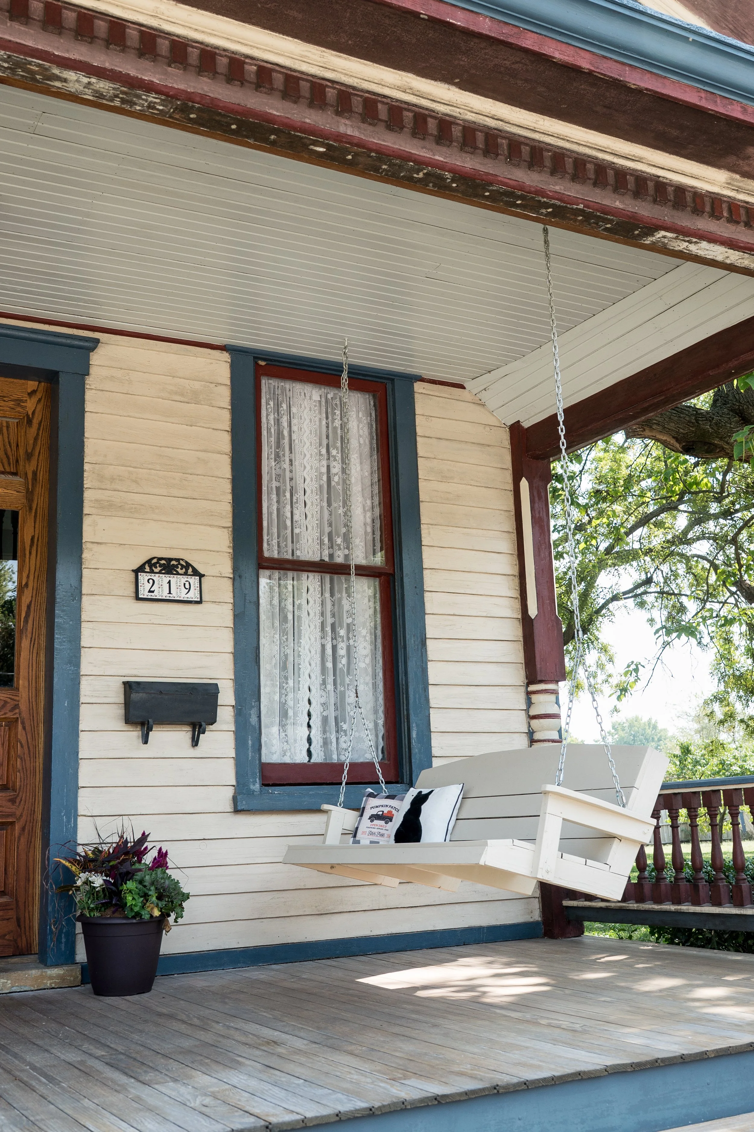 Porch with a white wooden swing, window with lace curtains, black mailbox, house number 219, flower pot, and attached railing, with trees in the background.