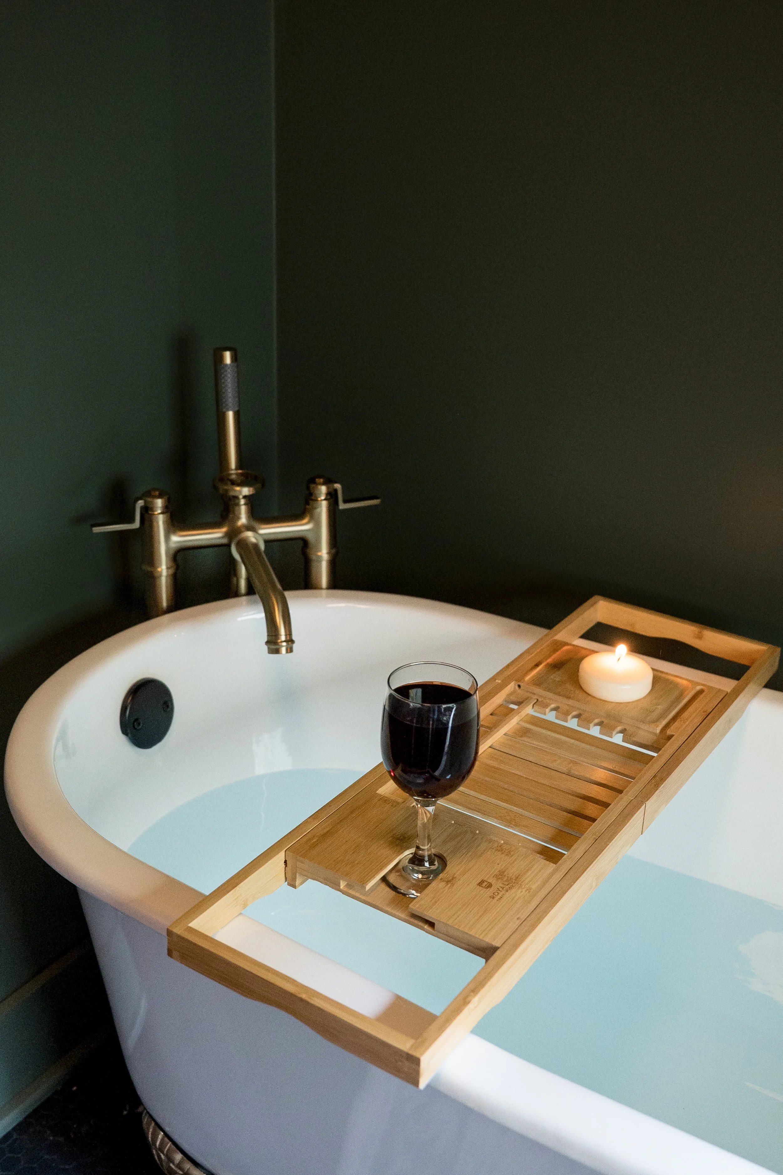 A bathtub with a wooden bath tray holding a glass of red wine, a lit candle, and a soap dish.