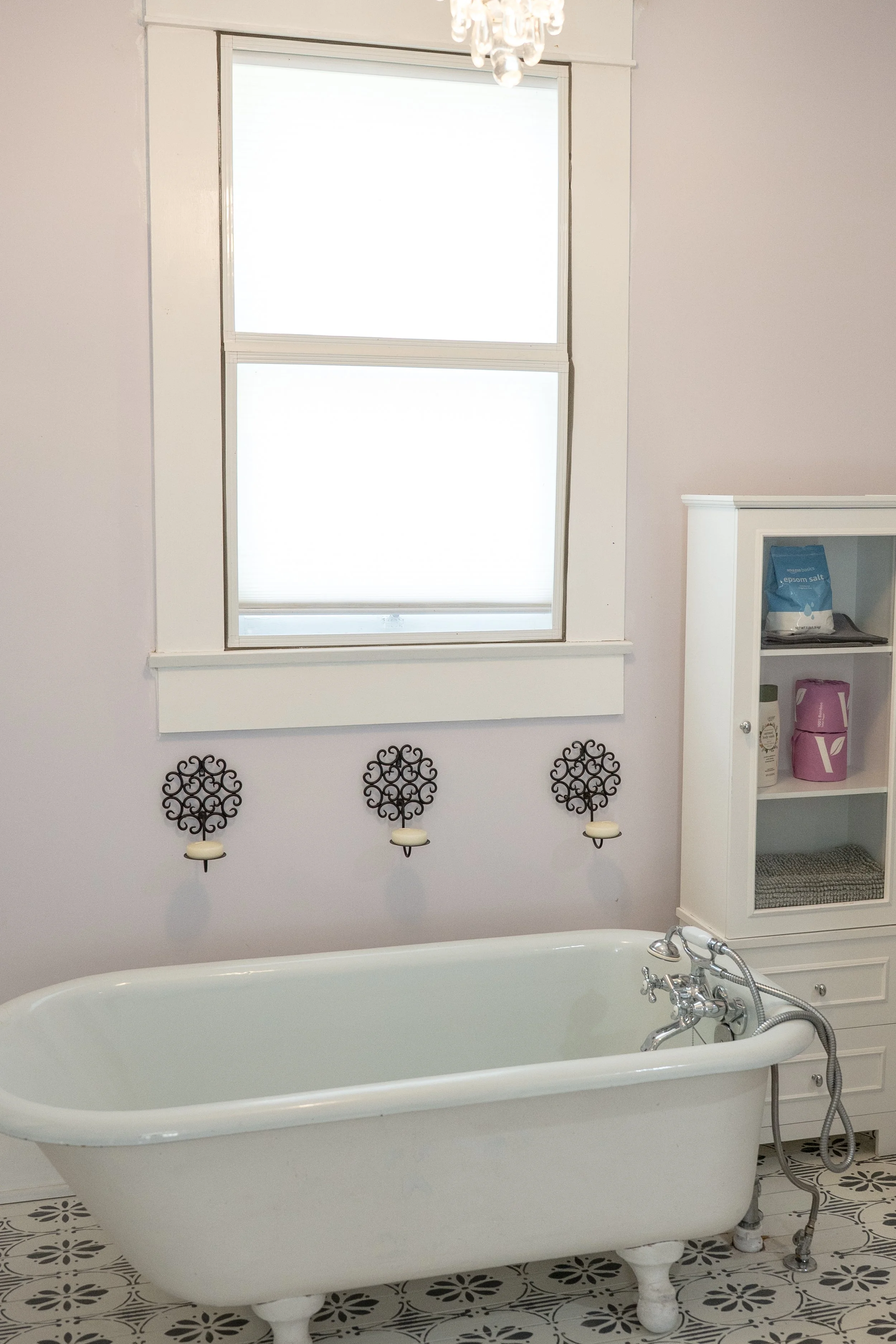 A vintage white clawfoot bathtub in a bathroom with patterned floor tiles, a window with blinds, and a white cabinet with toiletry items.