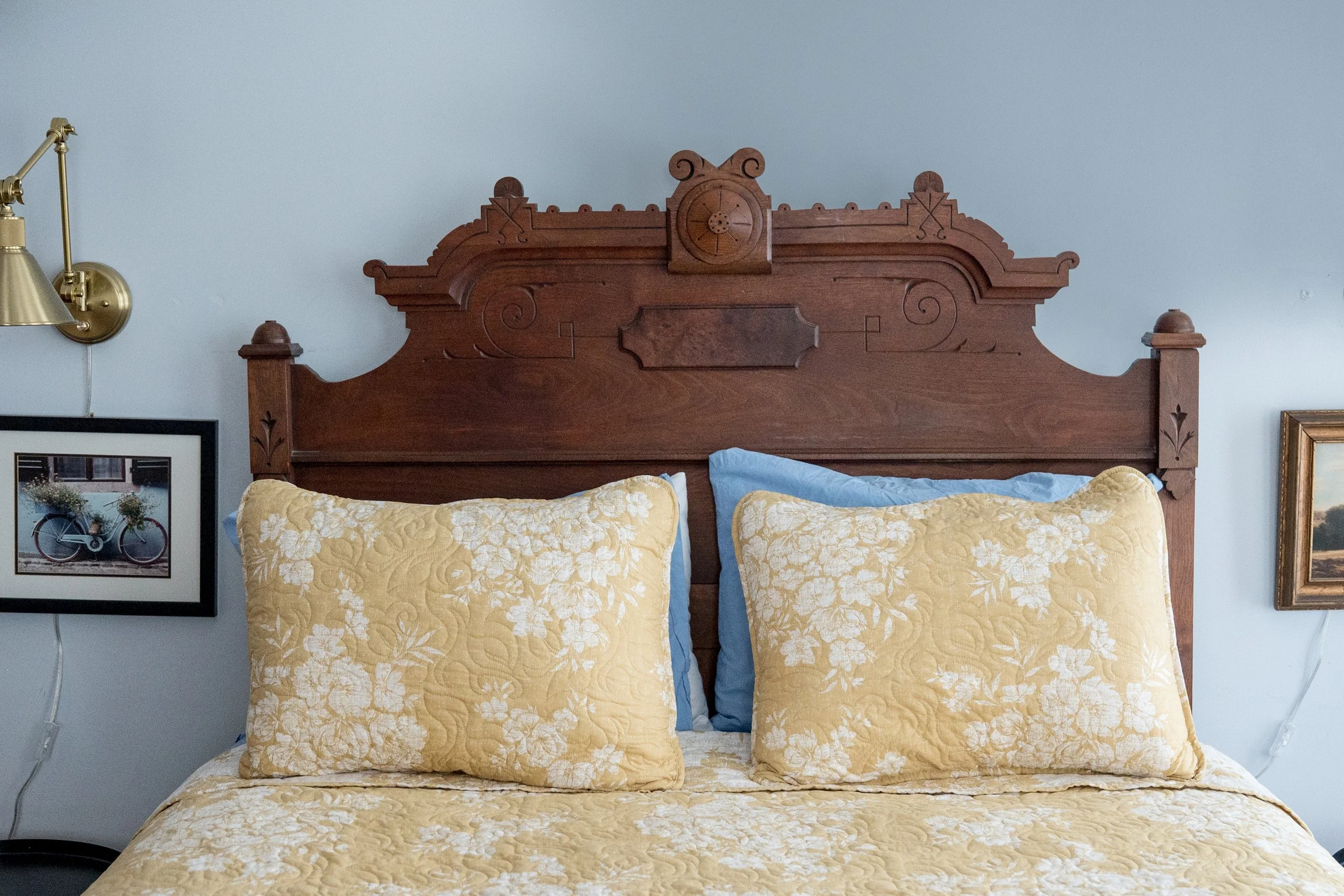 A bed with a wooden headboard, featuring a clock and decorative carvings, two large yellow pillows with white floral patterns, and two blue pillows, with framed pictures on the light blue wall beside it.