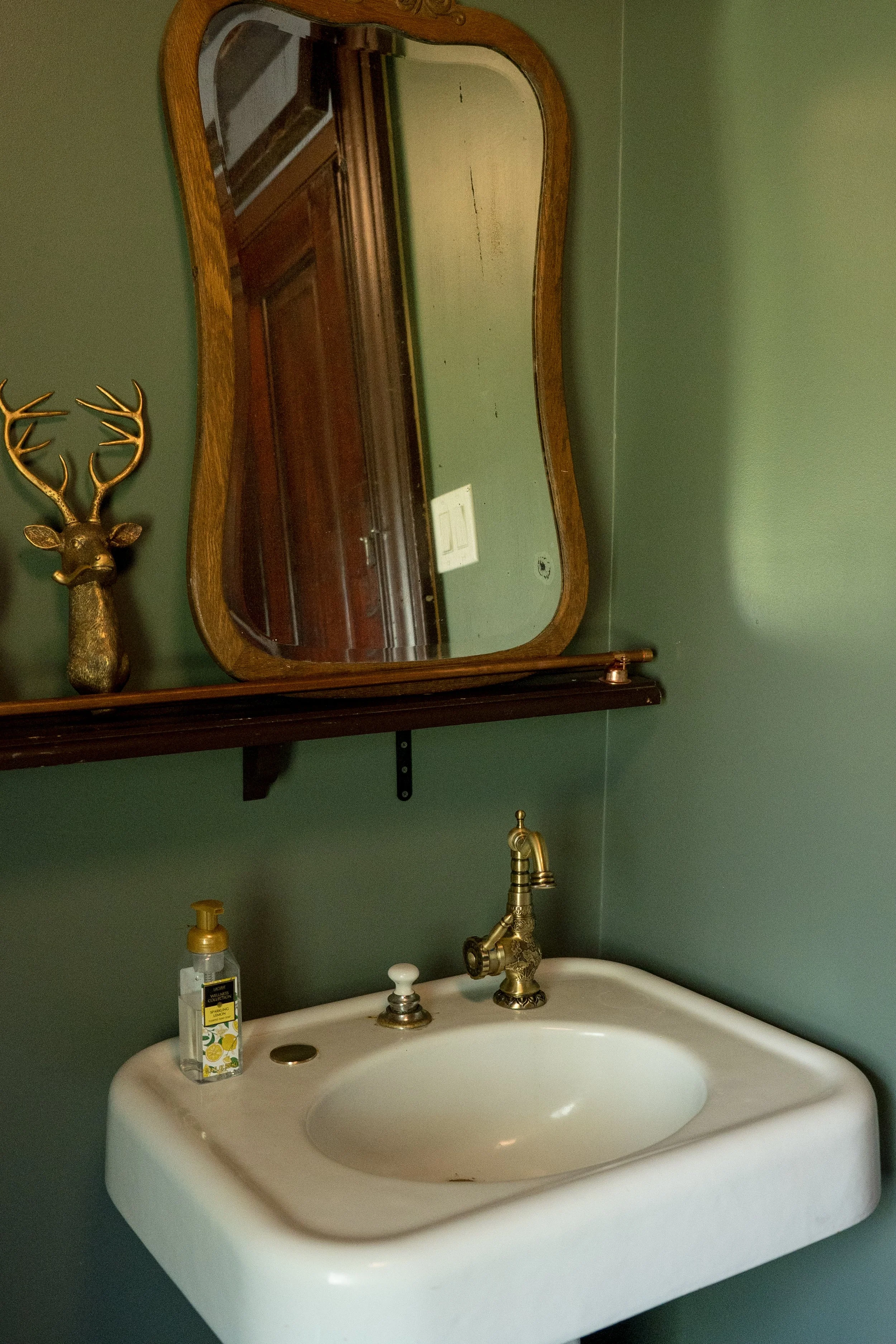 A bathroom sink with a gold faucet, a bottle of hand soap, and a mirror above it. The mirror has a wooden frame, and a decorative gold deer head is mounted on the wall to the left of the mirror. The sink and countertop are white, and the wall behind is painted green.