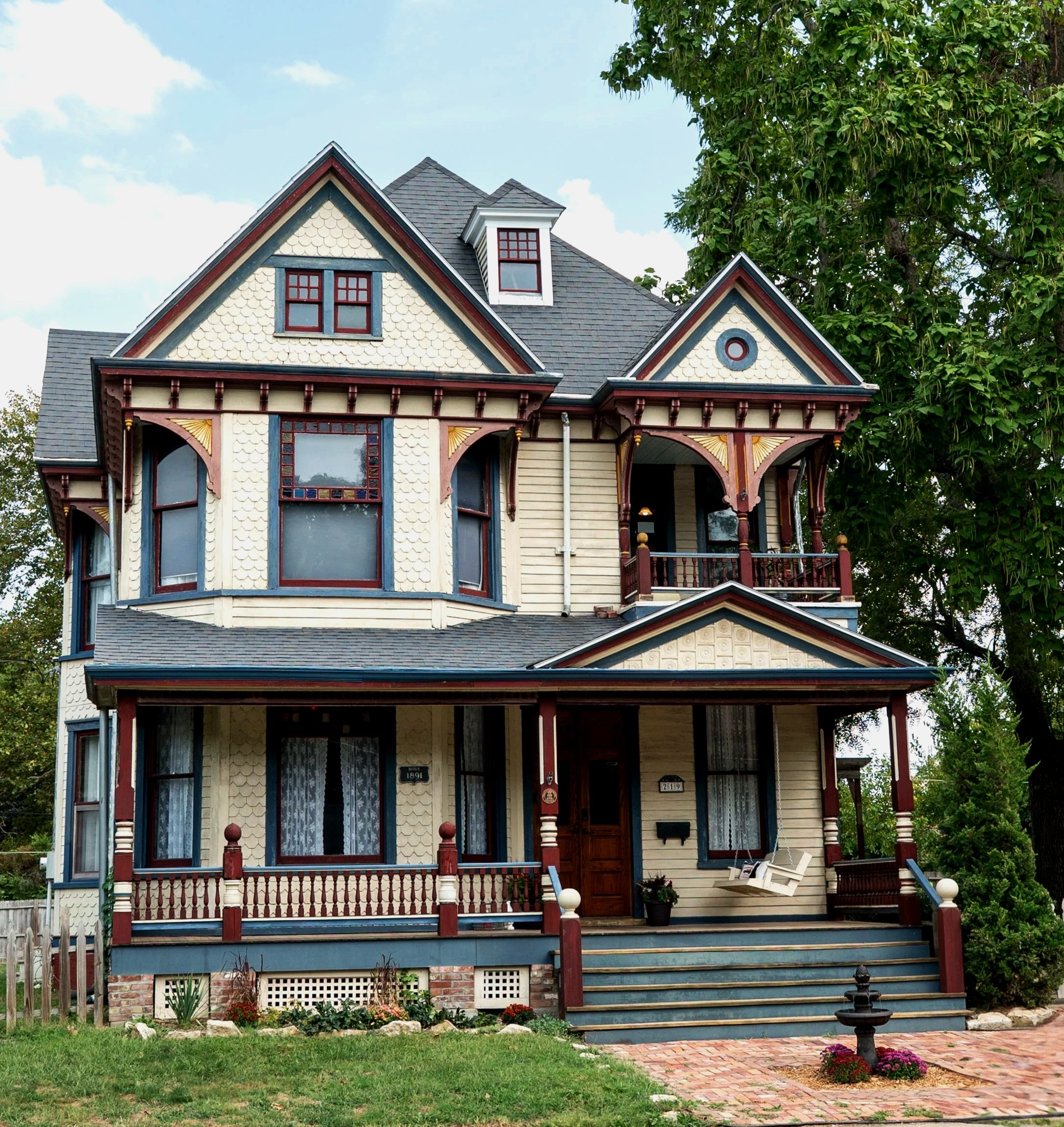 Victorian-style house with ornate trim, multiple gables, and a front porch with chairs and decorations, surrounded by a small garden and trees.