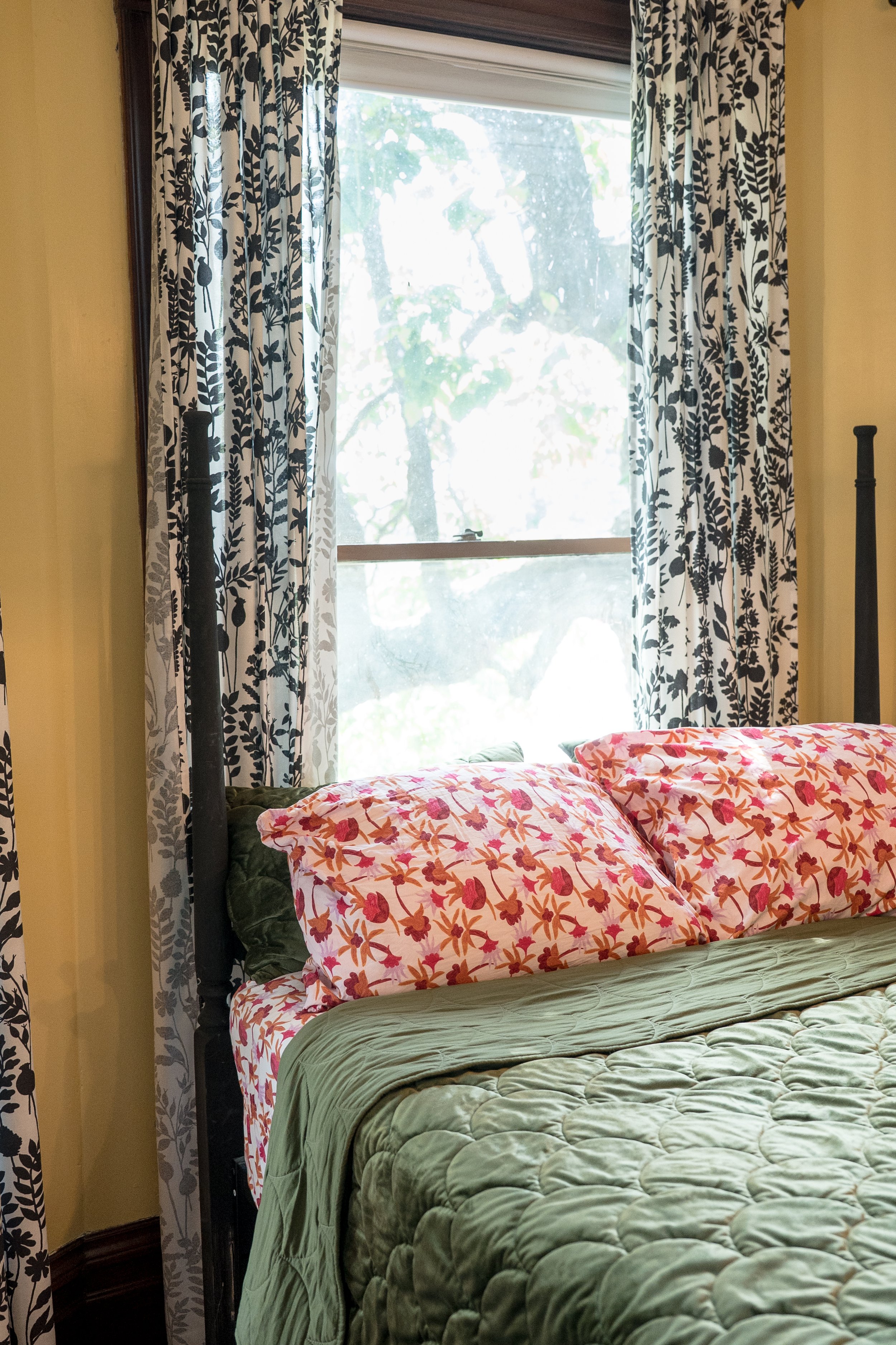A bedroom with a window and floral curtains, showing a bed with pink, orange, and yellow floral-patterned pillows, a green quilted bedspread, and a green blanket, with a tree visible outside the window.