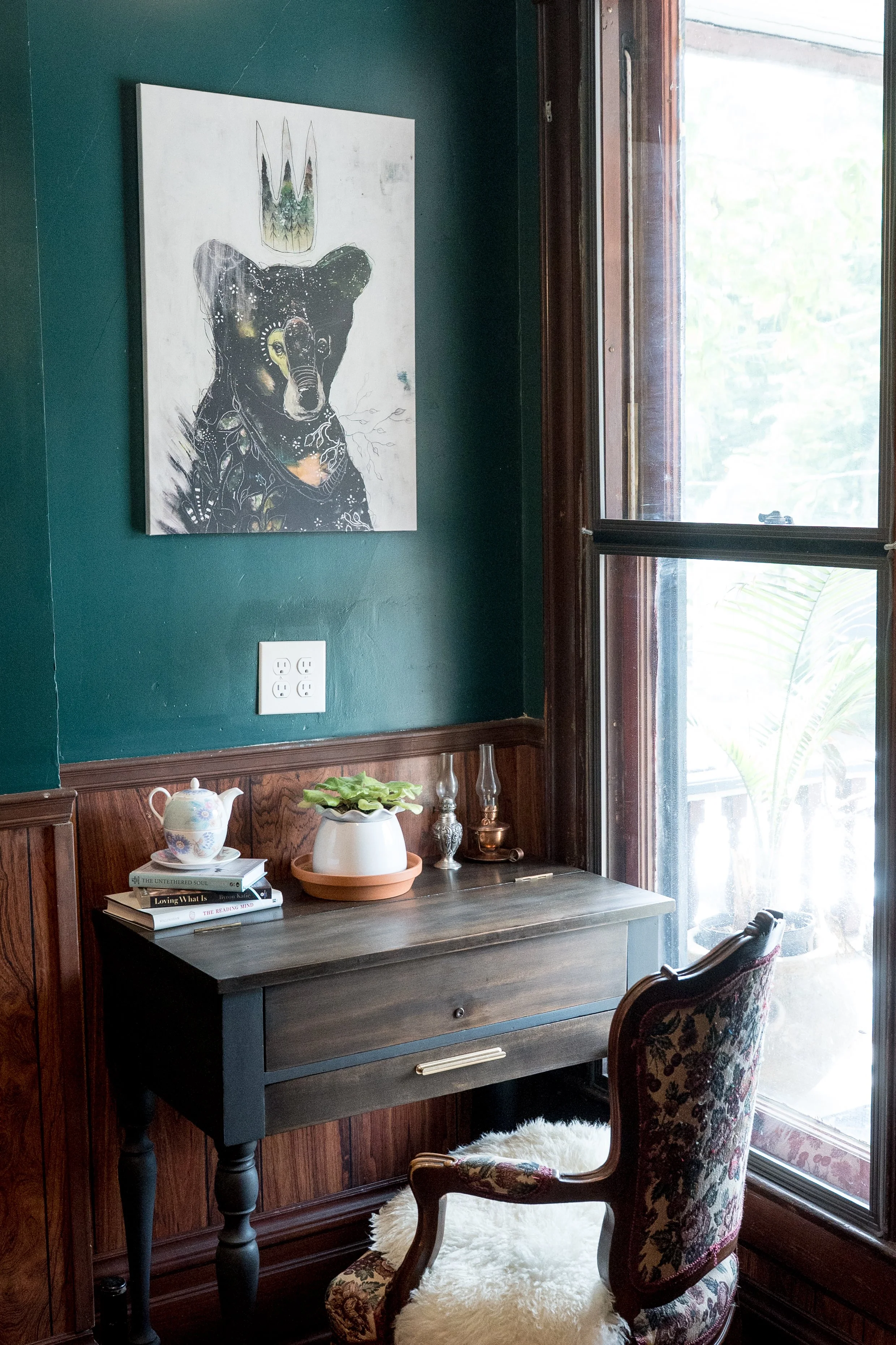 A cozy corner with a dark wooden desk, a vintage floral armchair, and a wall with dark green paint. The desk has a stack of books, a decorative teapot, a potted plant, and glass vases. A large window shows outdoor greenery and lets in natural light. A modern art painting of a bear with a crown hangs on the wall.