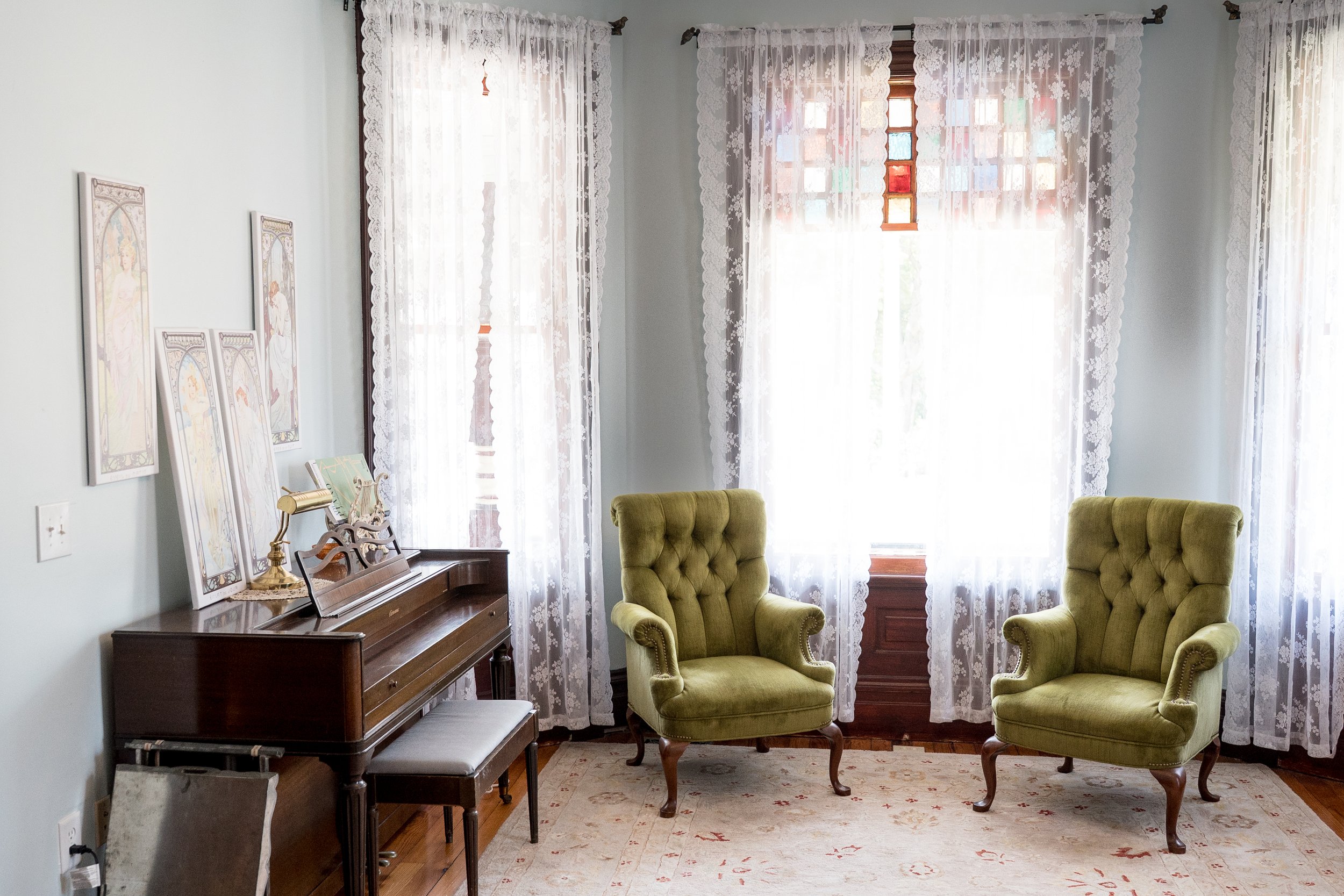 A cozy living room corner with vintage green upholstered armchairs, floral lace curtains on large windows, a wooden piano with decorative artwork leaning against the wall, and a plush rug on wooden floor.