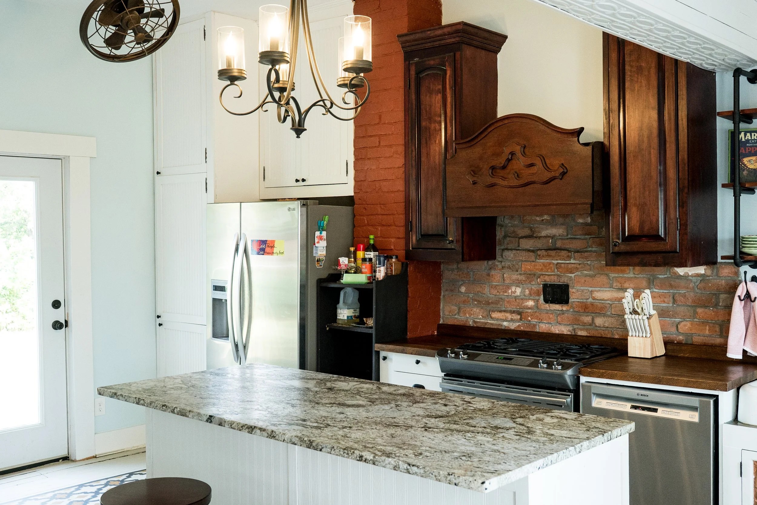 A kitchen with a large granite island, white cabinets, a stainless steel refrigerator, a gas stove, and a brick and wood accent wall with a wooden range hood. There is a chandelier hanging from the ceiling and some shelves on the brick wall.