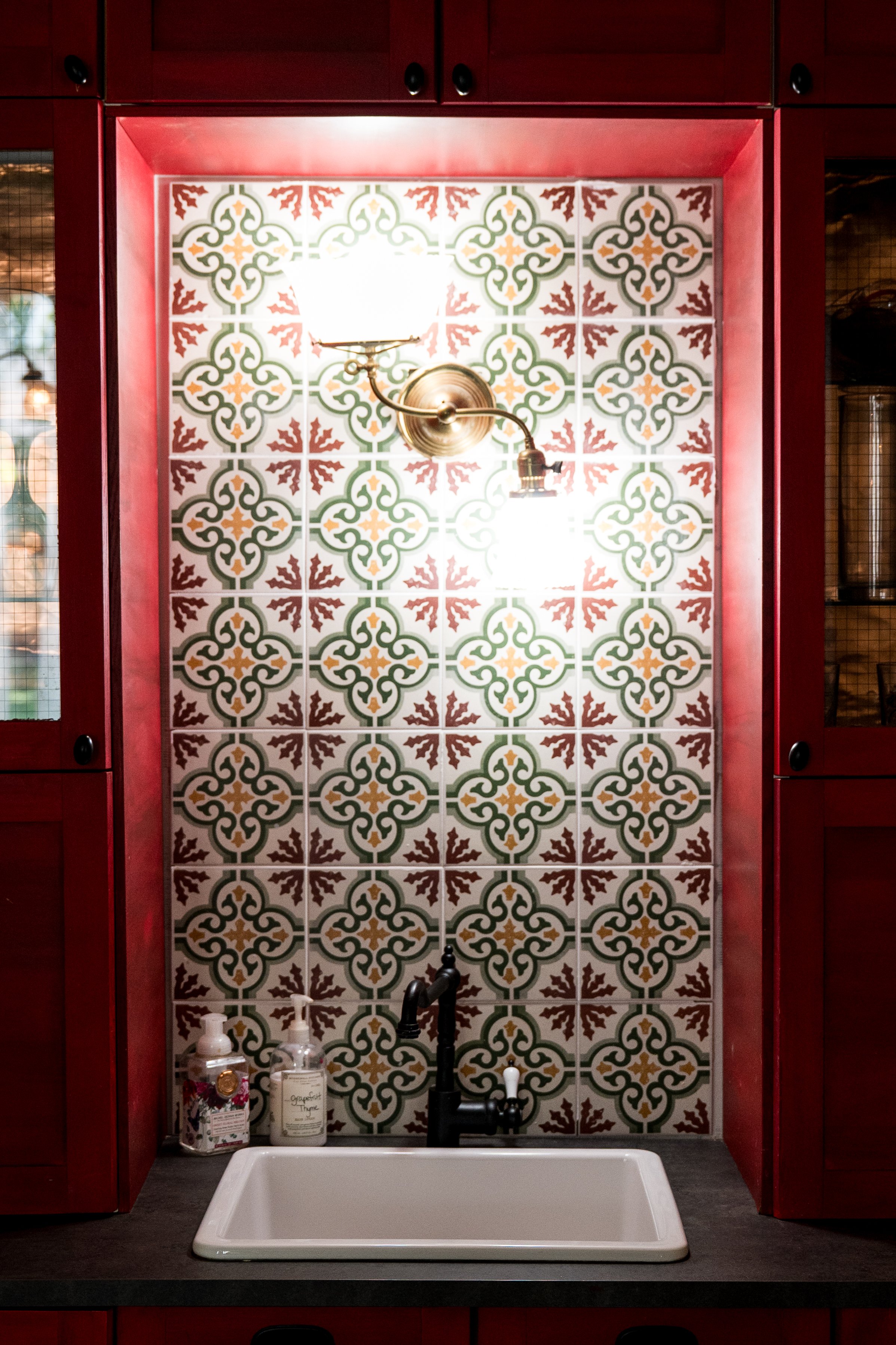 Kitchen wash station with a black faucet, white sink, patterned tile backsplash, red cabinetry, and a brass wall-mounted light fixture.