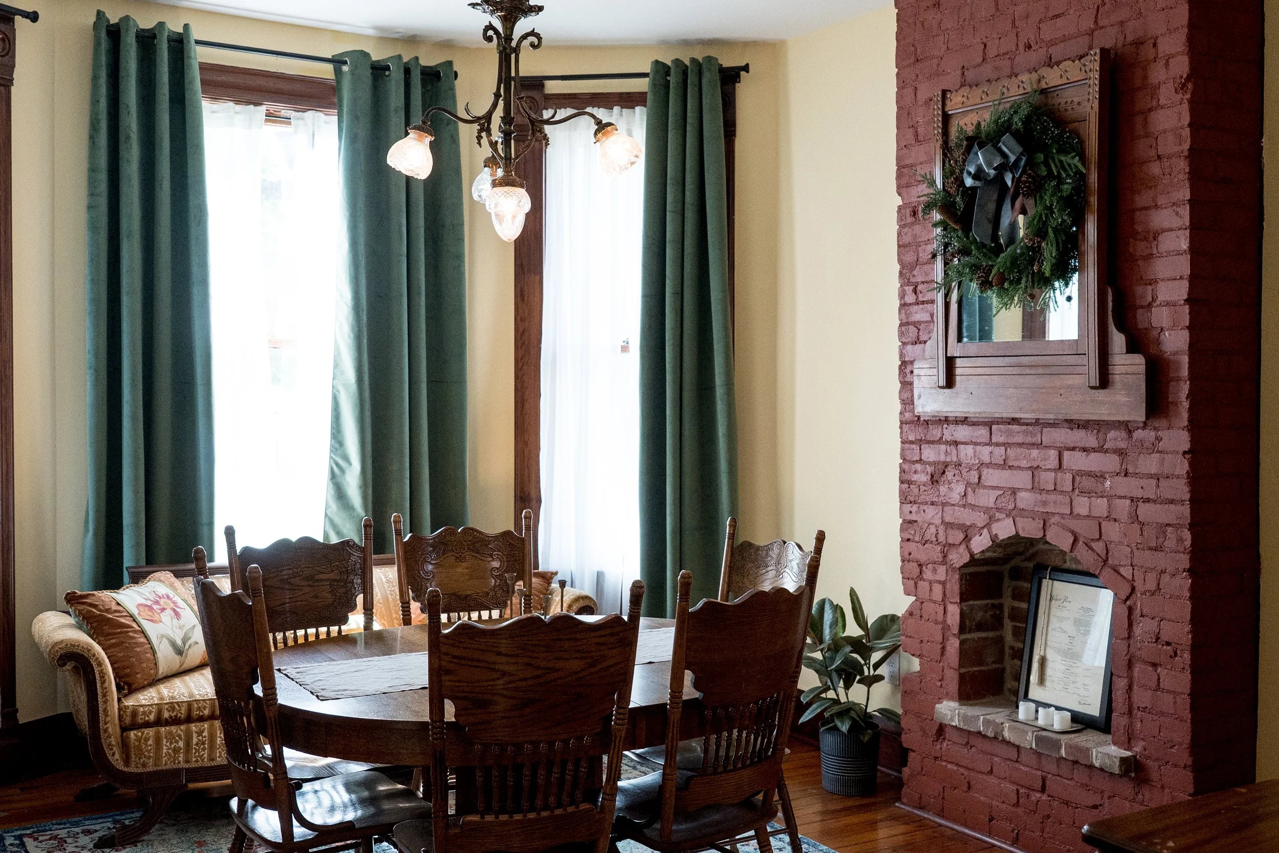 A dining room with a wooden table, chairs, and a floral couch. Large windows with green curtains and white sheer curtains. A chandelier hangs from the ceiling. On the red brick wall, there is a decorative mirror with greenery and a black ribbon. A framed certificate or artwork leans against the wall near a plant and candles.