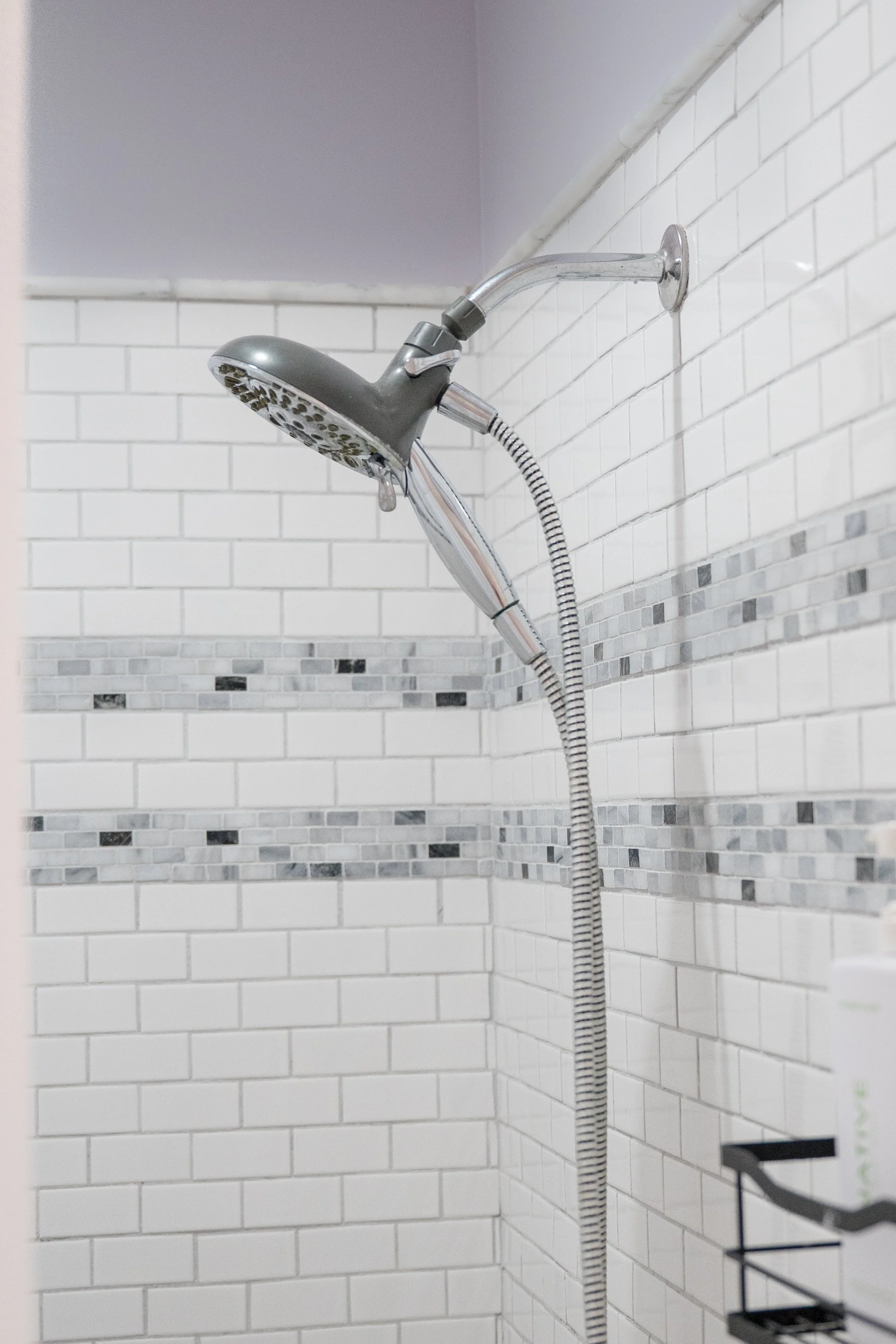 A showerhead and handheld shower hose attached to a tiled wall in a bathroom with white rectangular tiles and a strip of small mosaic tiles.
